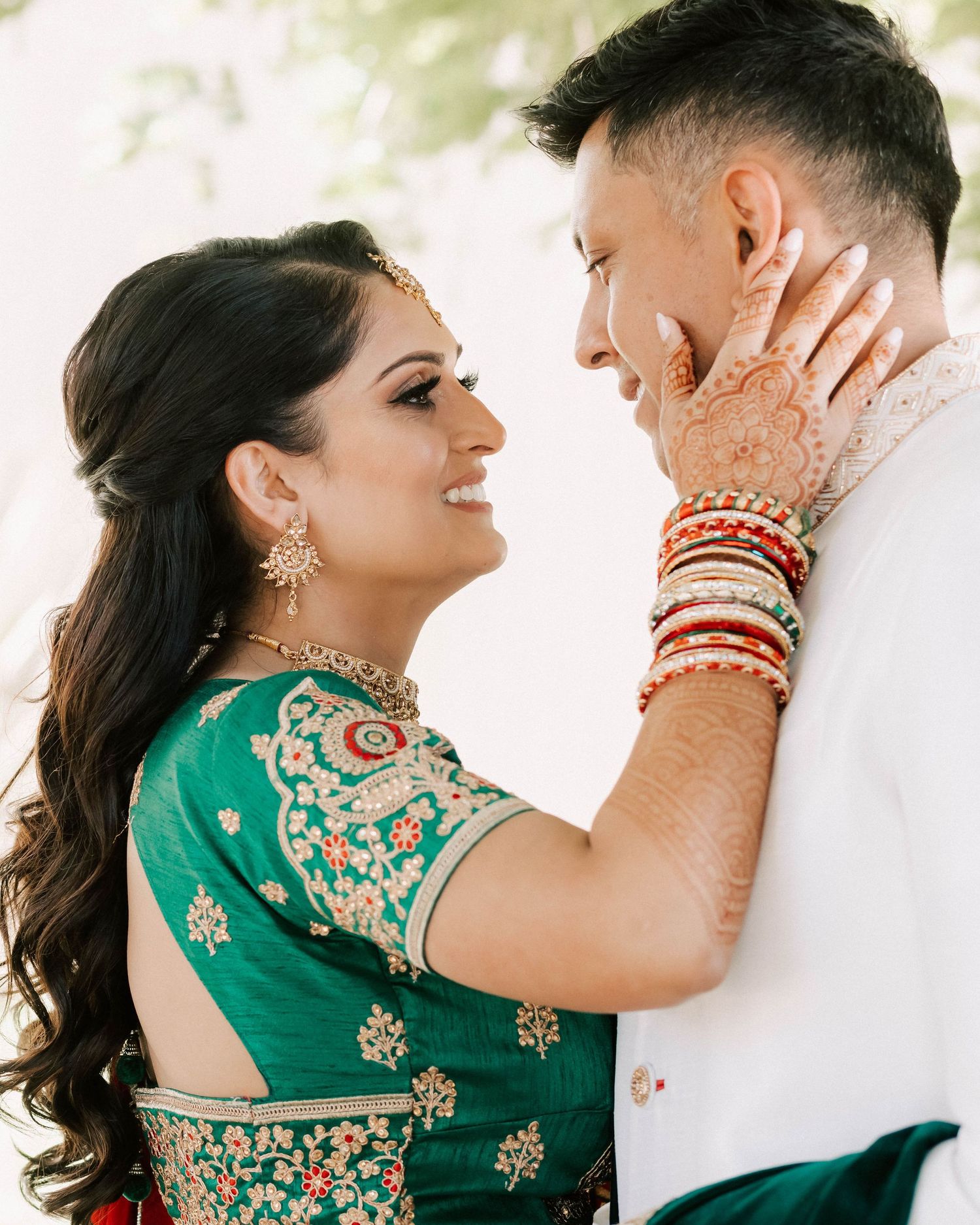 Indian bride in green lehenga with mehndi hands tenderly touching grooms face outdoors.