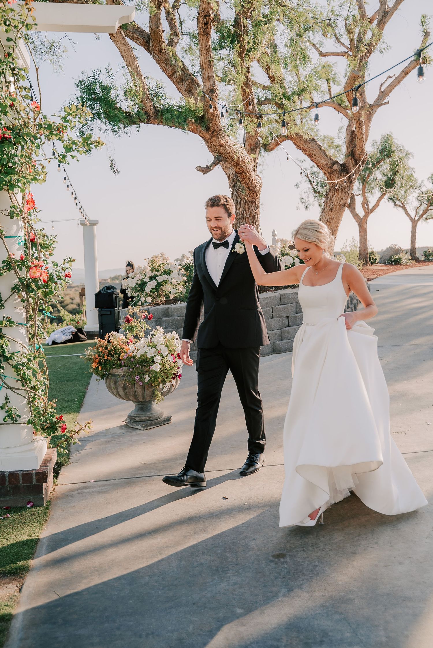 Groom in black tuxedo twirls bride in white ball gown outdoors near floral arch and trees at elegant wedding venue.