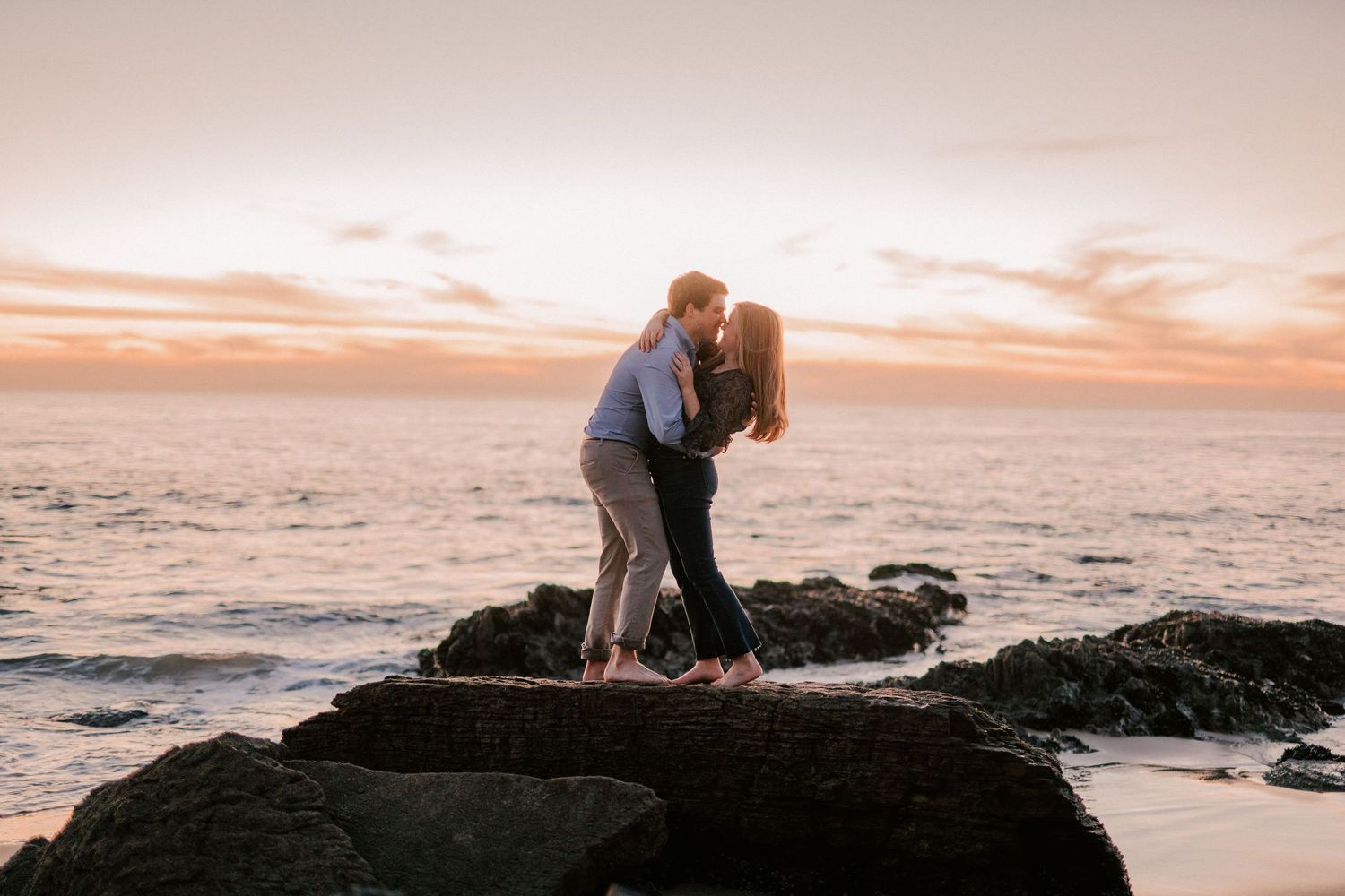 Couple sharing a romantic kiss on rocky shoreline during golden sunset over the ocean.