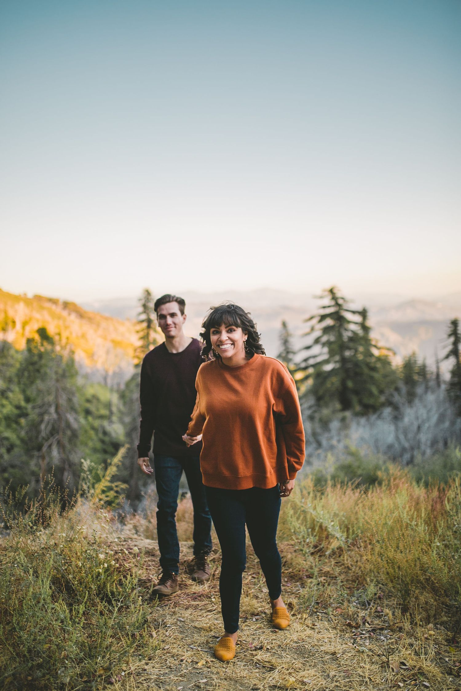 Smiling couple holding hands on a mountain trail at sunset with scenic views.