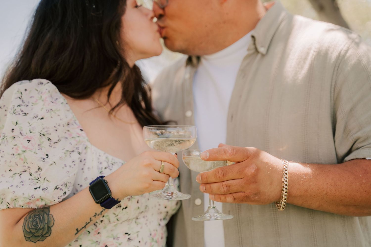 Couple kissing and clinking champagne glasses outdoors, woman with tattoo and floral dress, man in beige shirt.