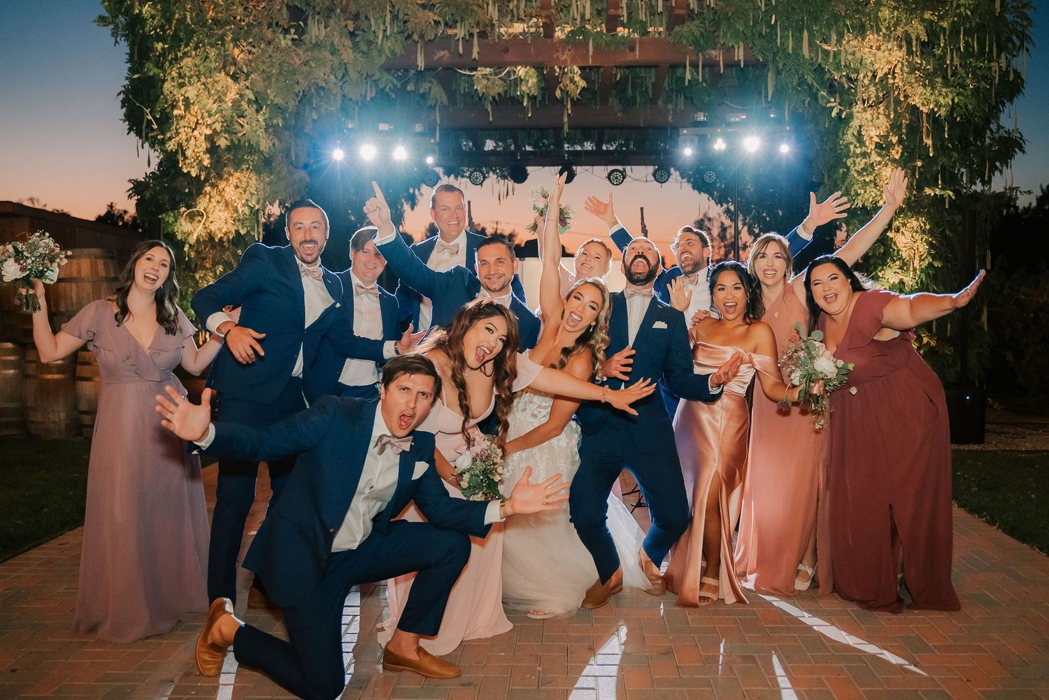Wedding party celebrating outdoors at dusk, bride and groom surrounded by bridesmaids and groomsmen in navy suits.