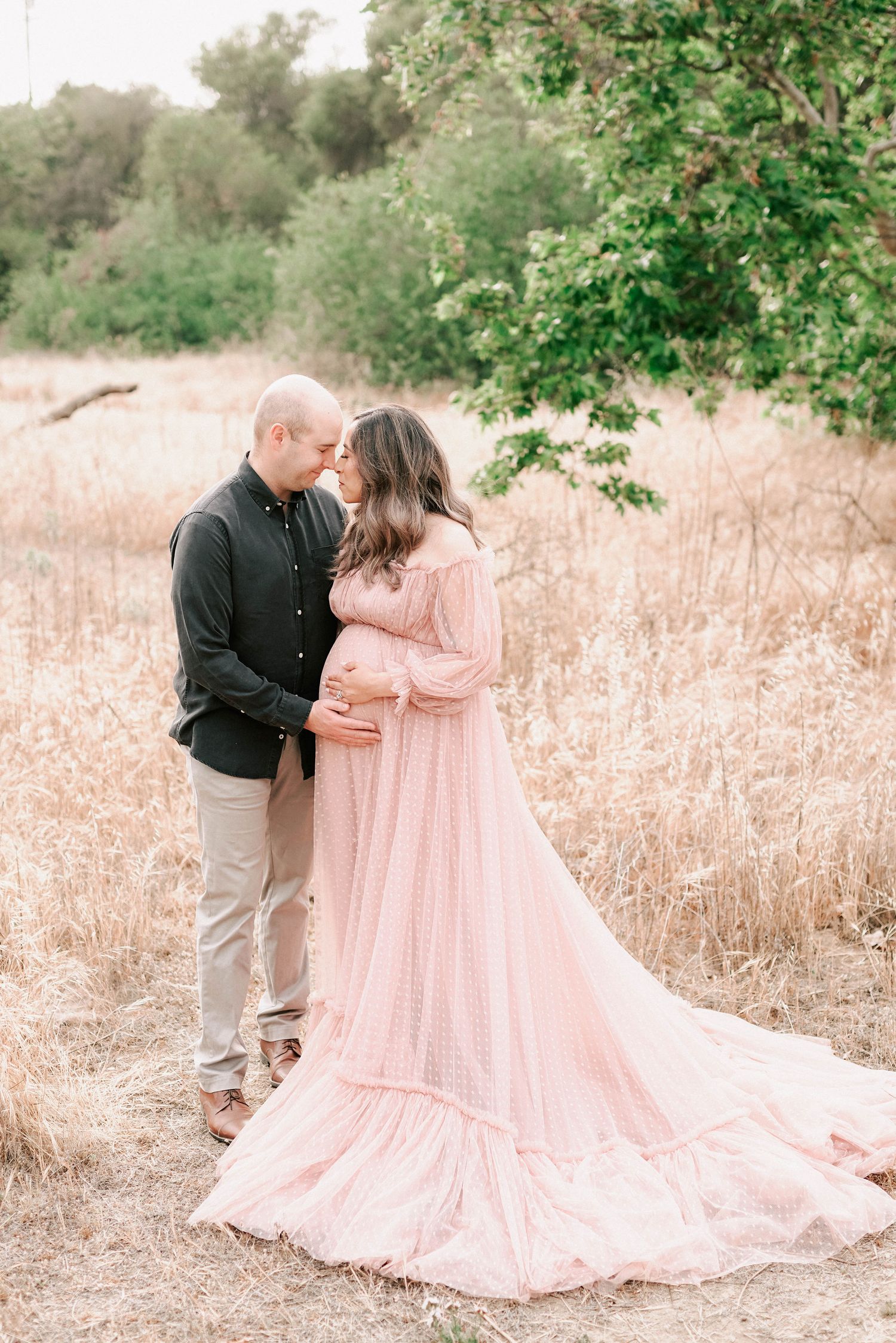 Couple in a field, pregnant woman in flowing pink gown, man in black shirt, sharing tender moment.