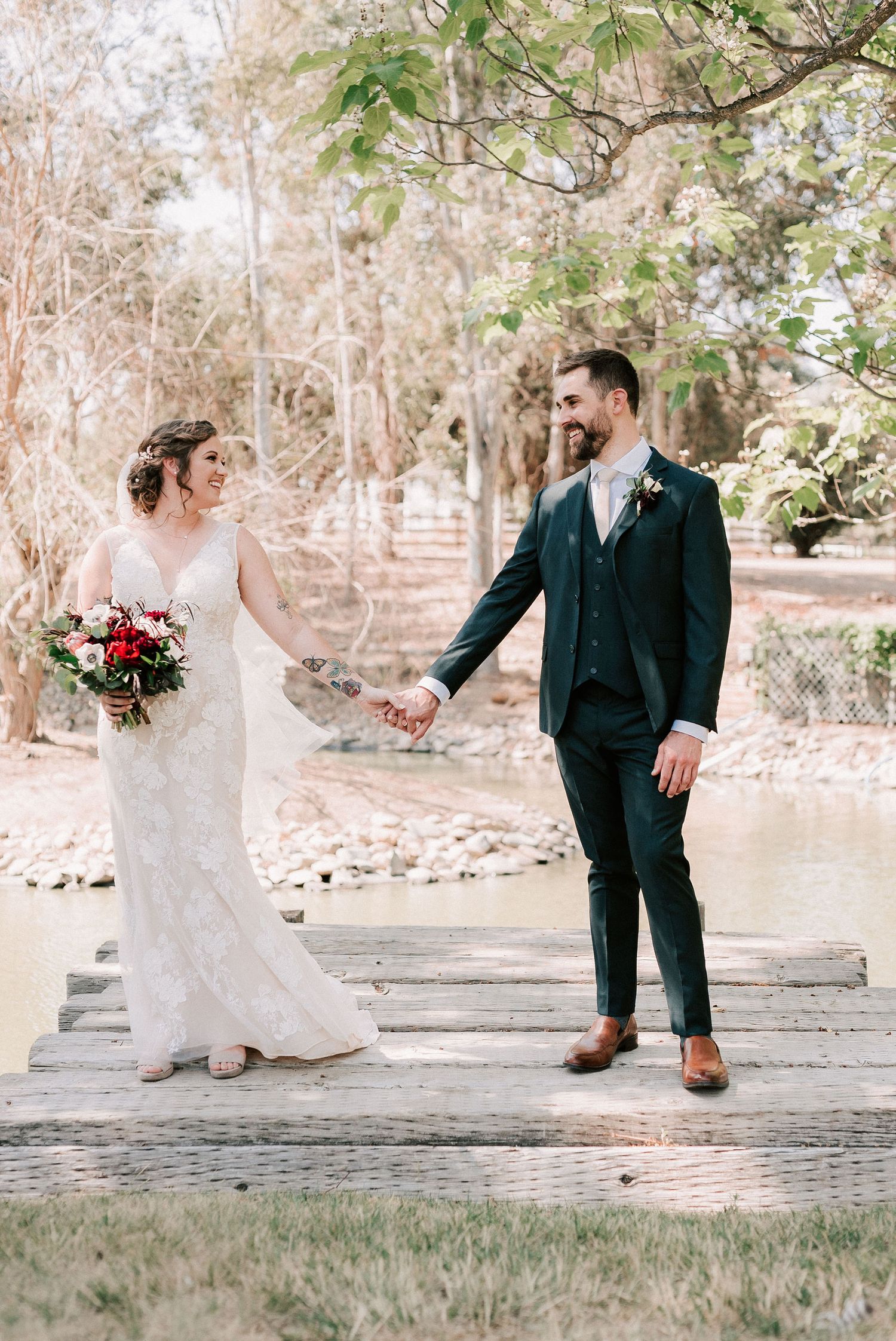 Bride and groom hold hands on a wooden dock surrounded by trees, she holds a red bouquet, he wears a dark suit.
