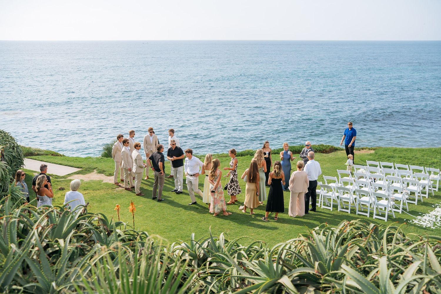 Wedding guests mingle on a lush green oceanfront lawn with white chairs after an outdoor seaside ceremony.