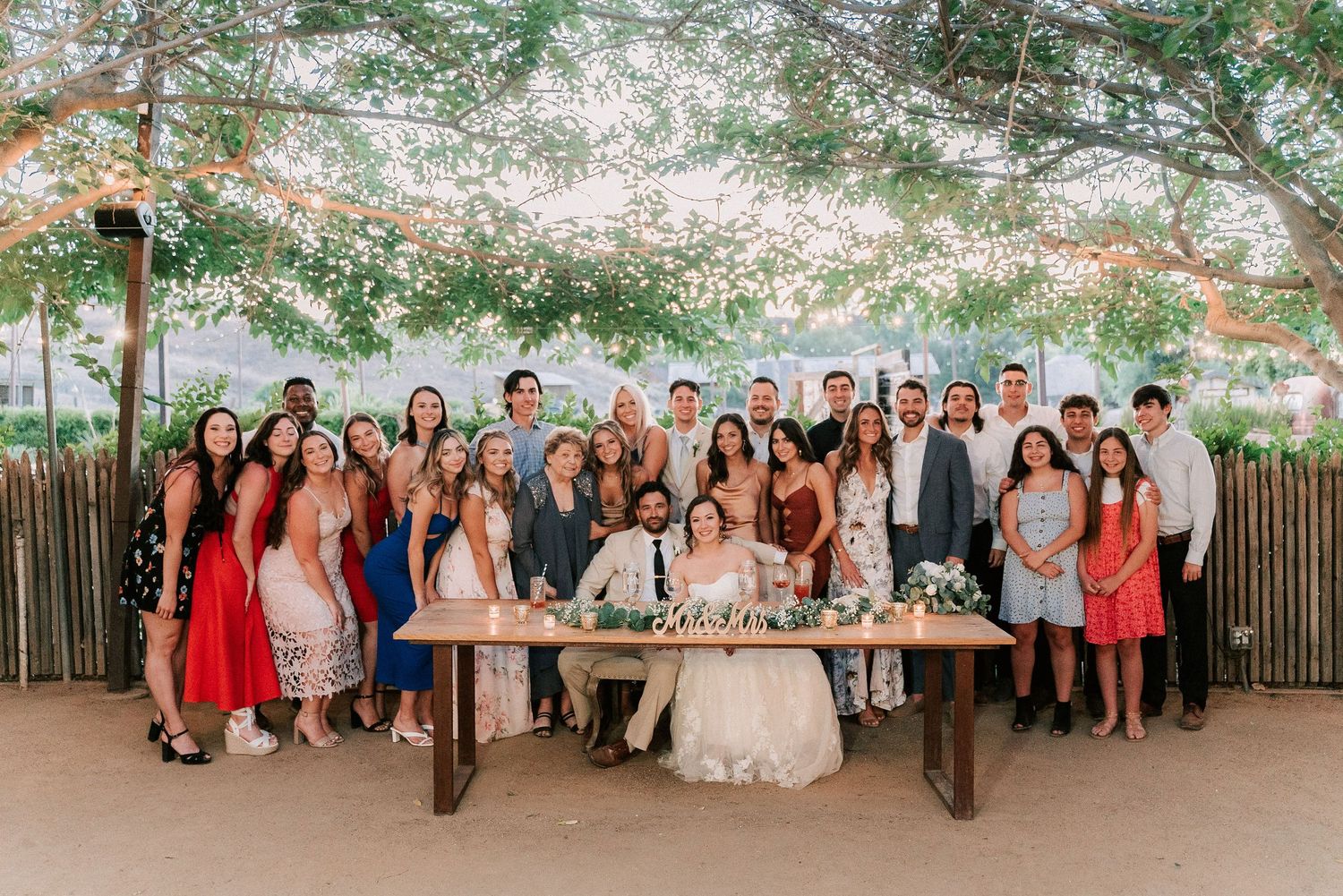 Wedding party poses with bride and groom at reception table under string lights and lush green canopy.
