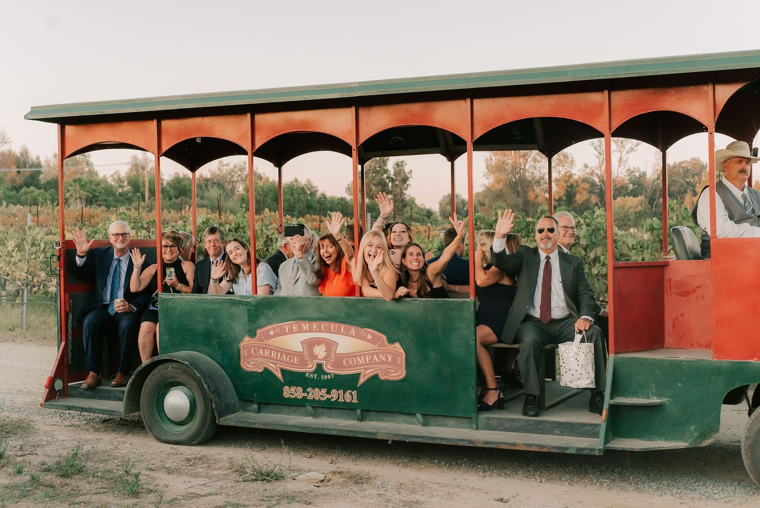 Group of cheerful wedding guests waving from a vintage red and green trolley at an outdoor orchard venue.