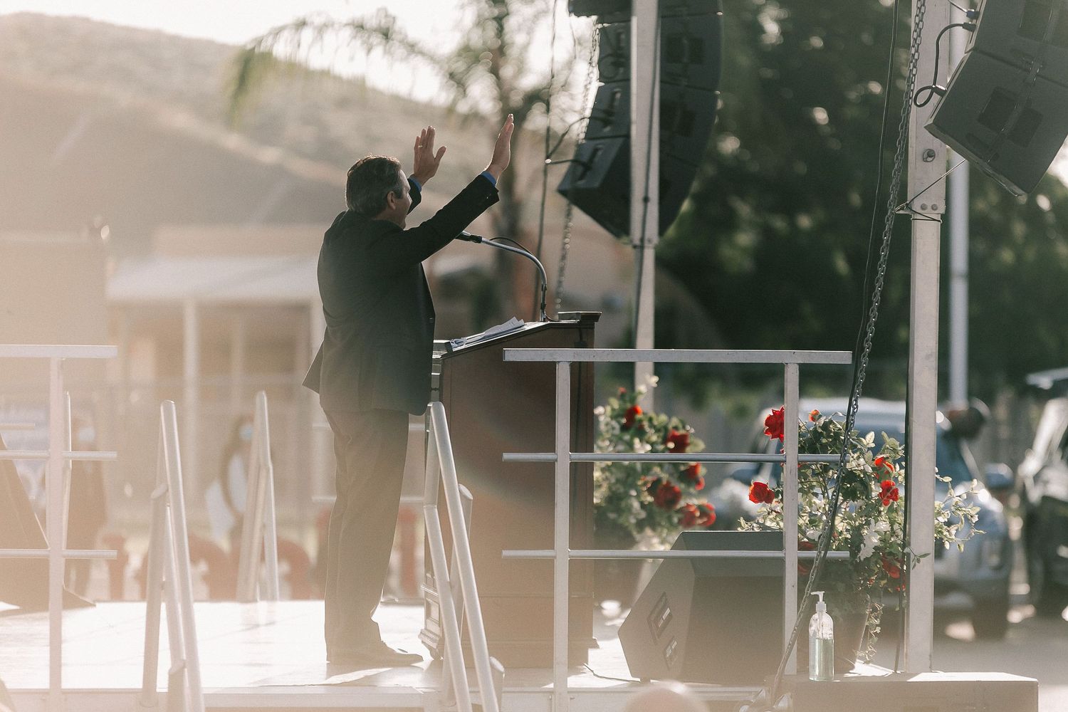 A speaker at a podium gestures on an outdoor stage adorned with a floral wreath in warm sunlight.