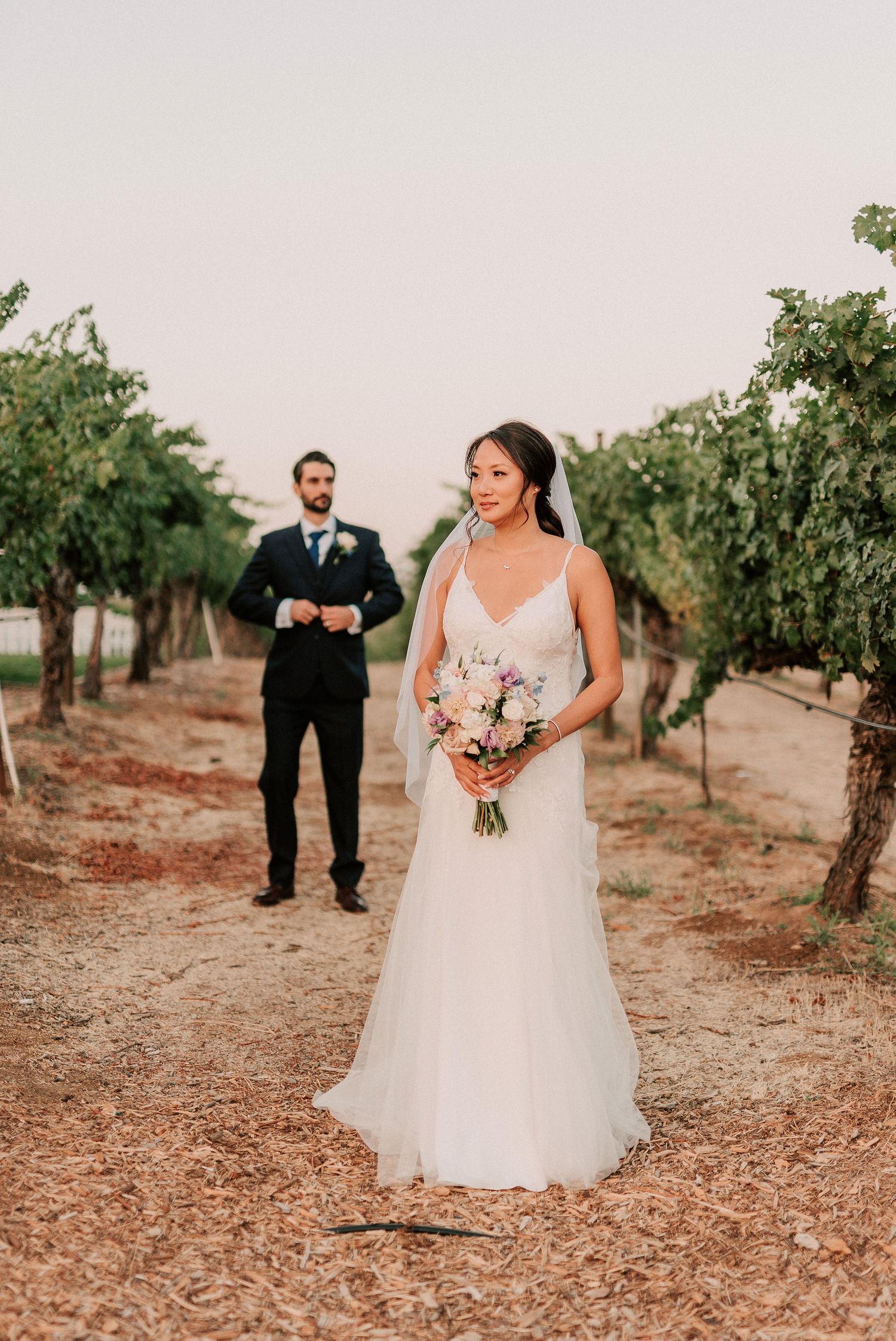 Bride in white gown holding bouquet stands in vineyard with groom in black suit behind her during golden hour.