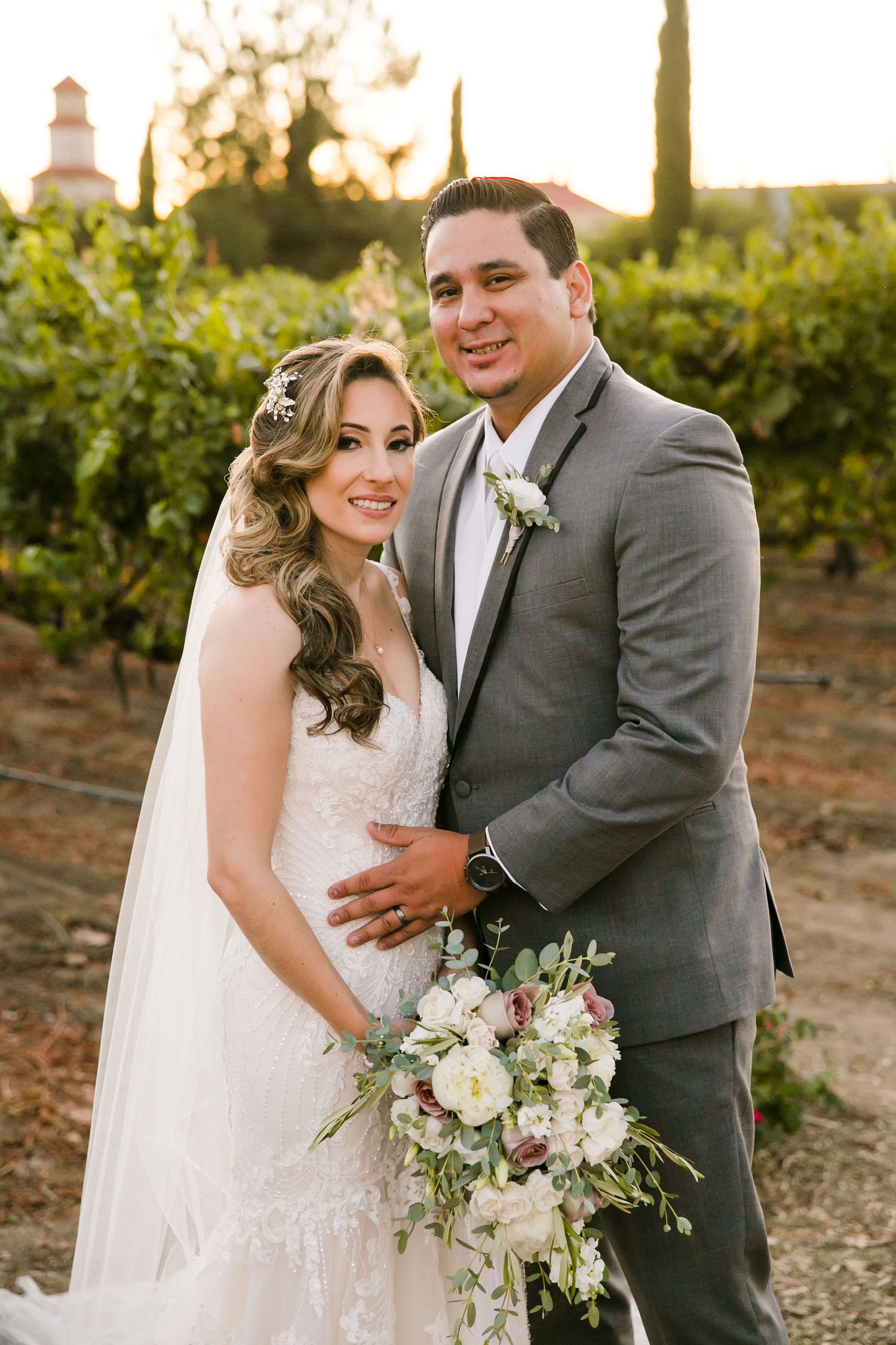 Bride and groom posing together in a vineyard at sunset, bride holding white and mauve floral bouquet.