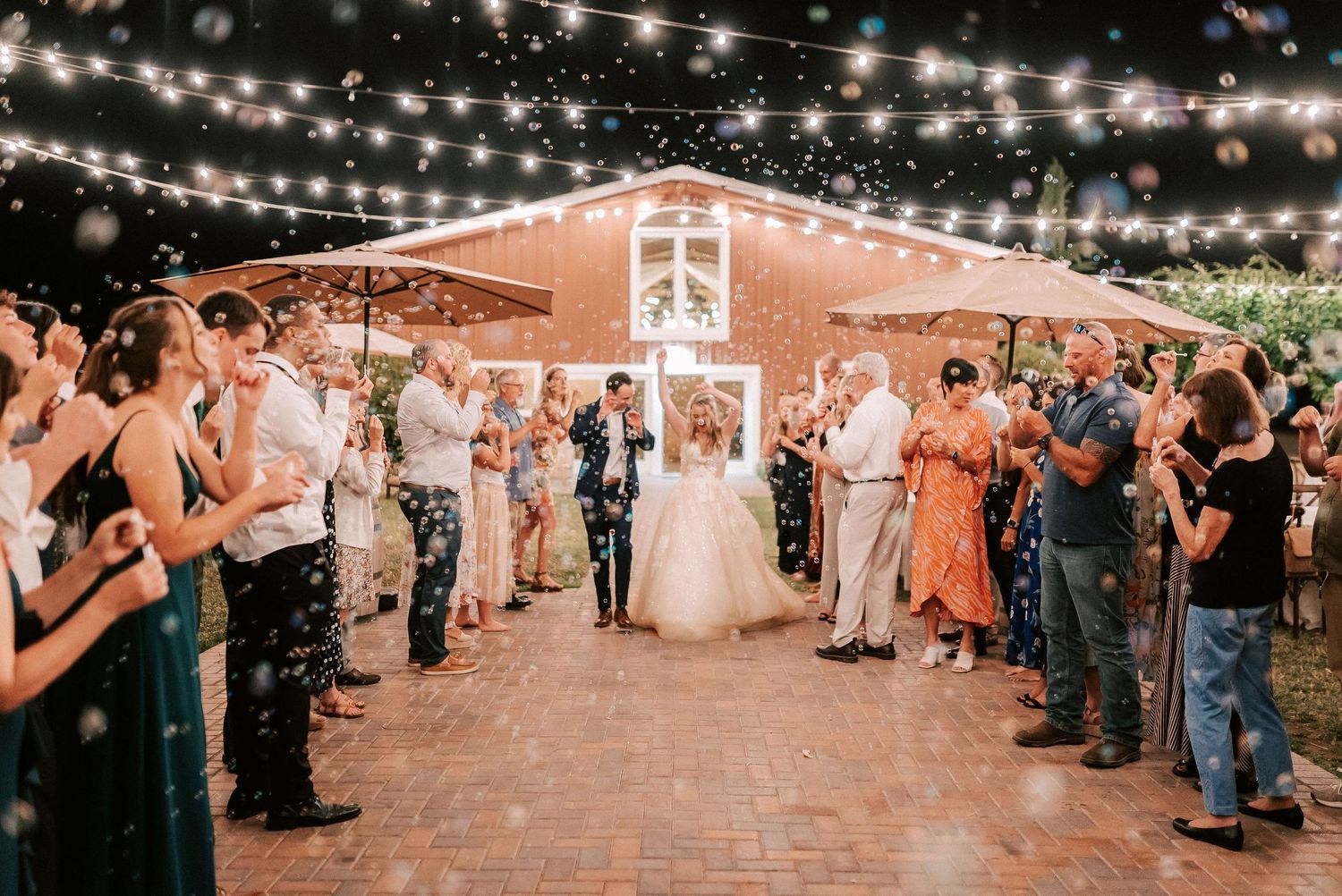 Bride and groom exit wedding reception under string lights with guests blowing bubbles at outdoor venue.