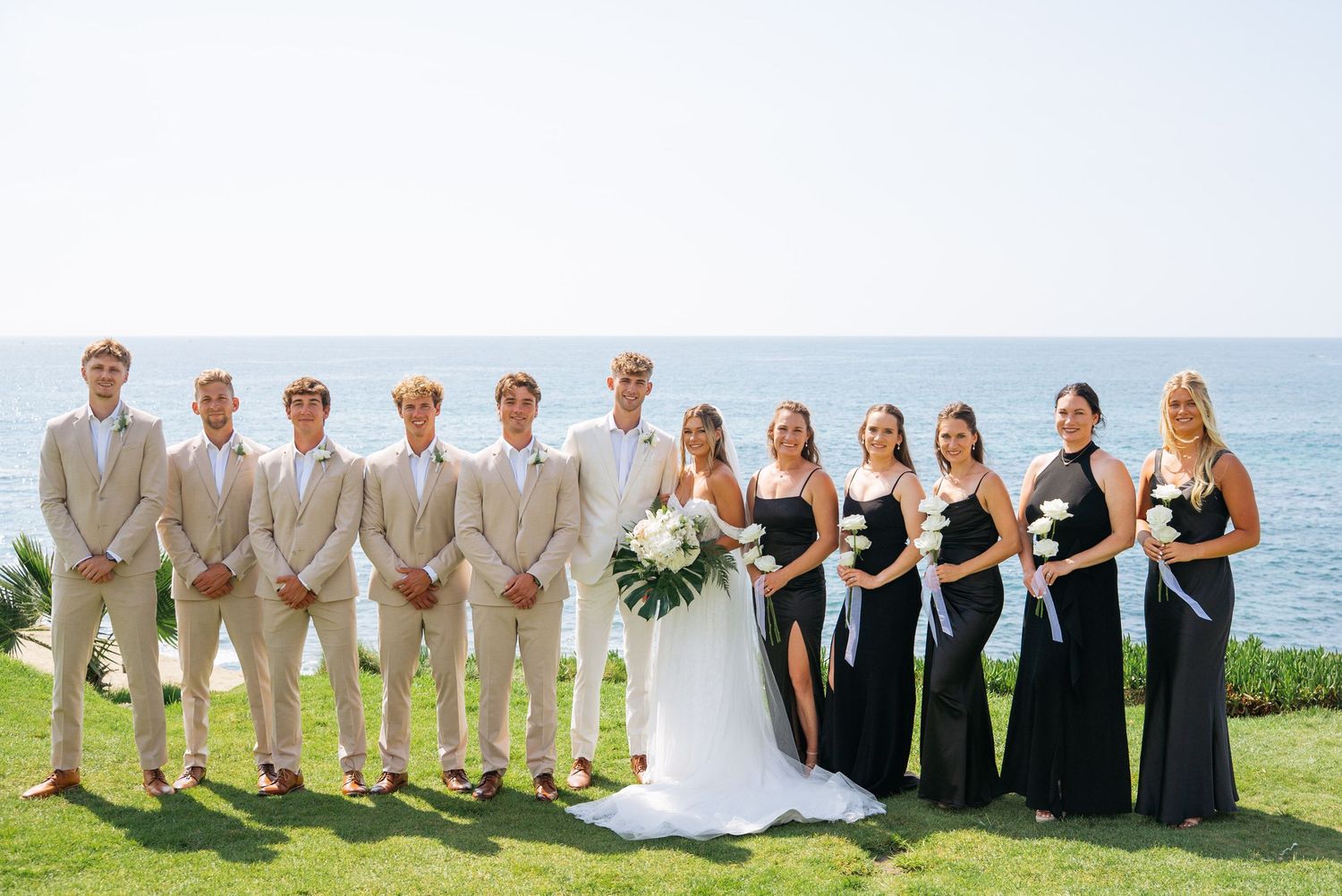 Wedding party poses outdoors by ocean, groomsmen in tan suits, bridesmaids in black gowns, bride in white dress.