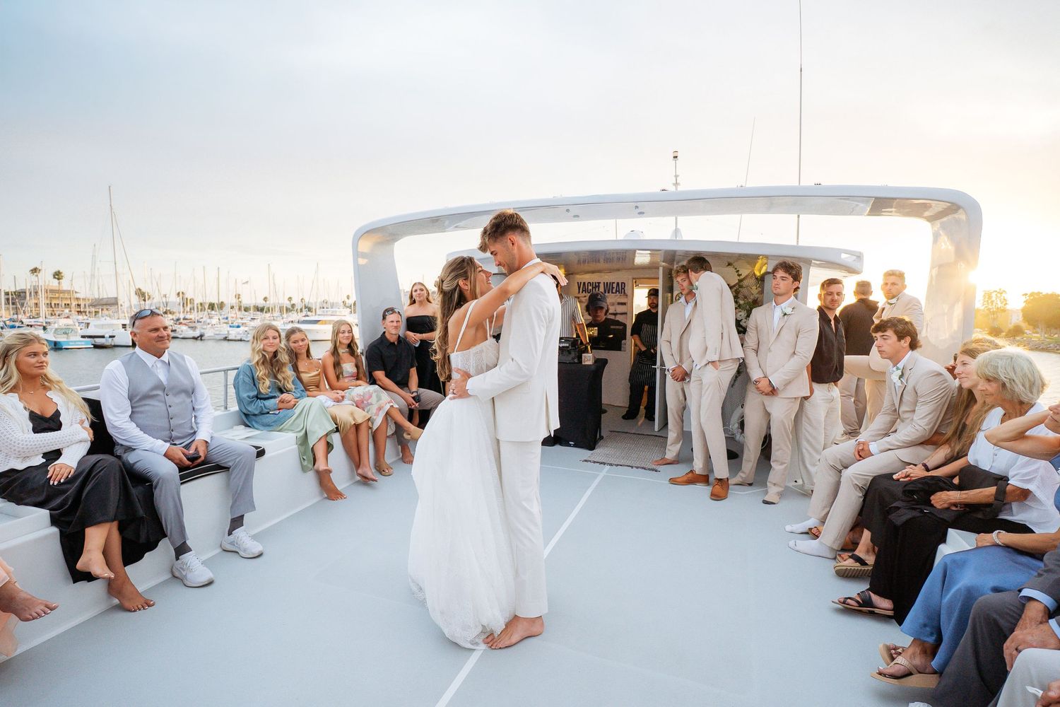 Bride and groom share first dance on yacht deck at sunset surrounded by seated wedding guests in a marina.