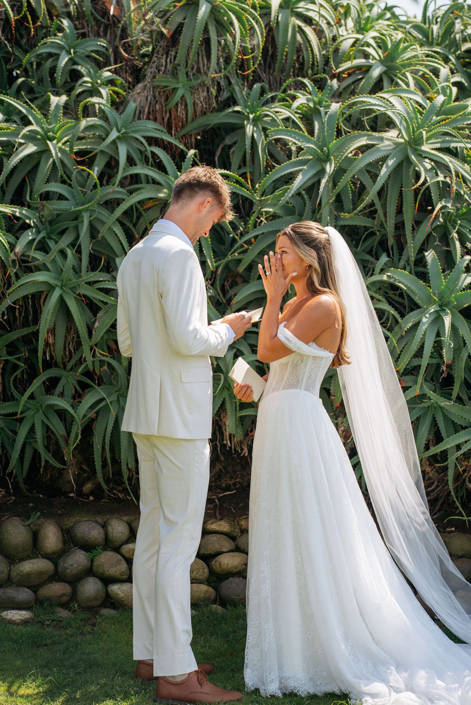 Emotional bride covers face as groom reads vows in lush tropical garden setting during outdoor wedding ceremony.