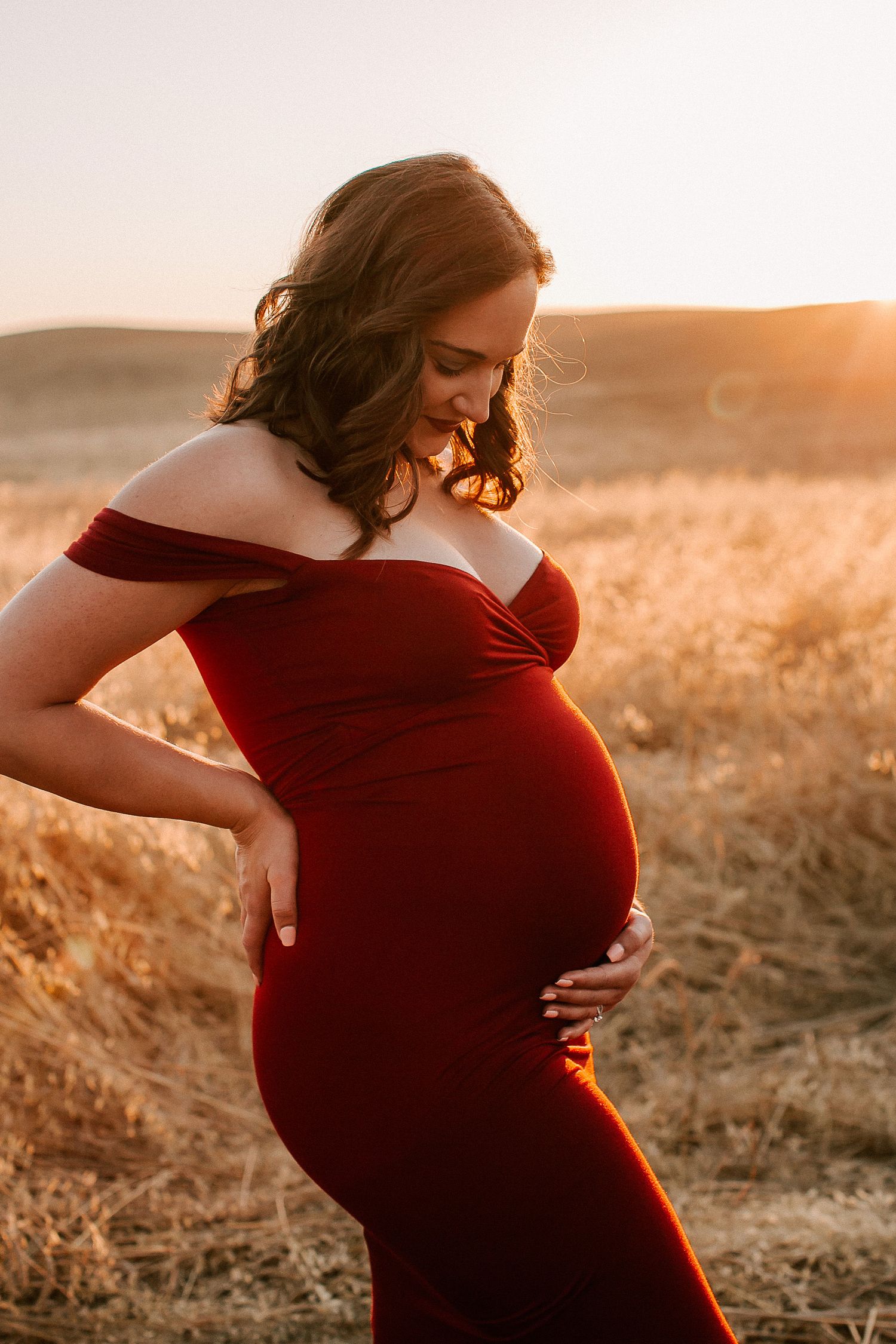 Pregnant woman in red off-shoulder gown cradling baby bump at golden sunset in a field.