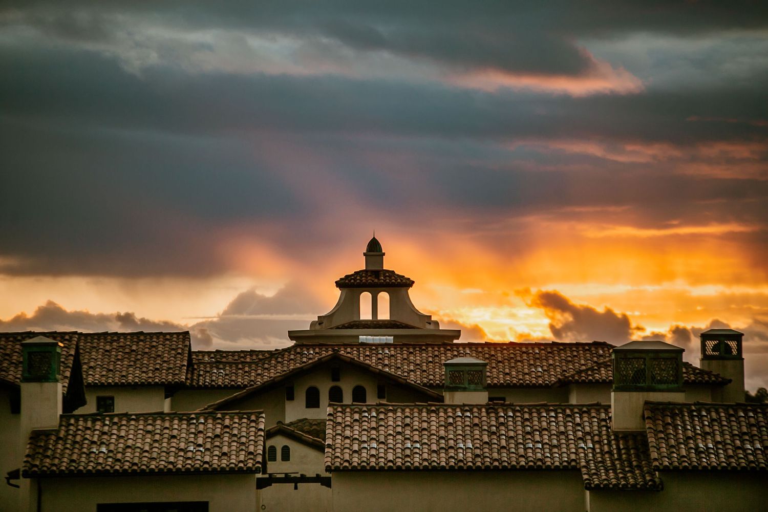 Dramatic sunset sky with golden rays behind historic tiled rooftops and a cupola under stormy clouds.