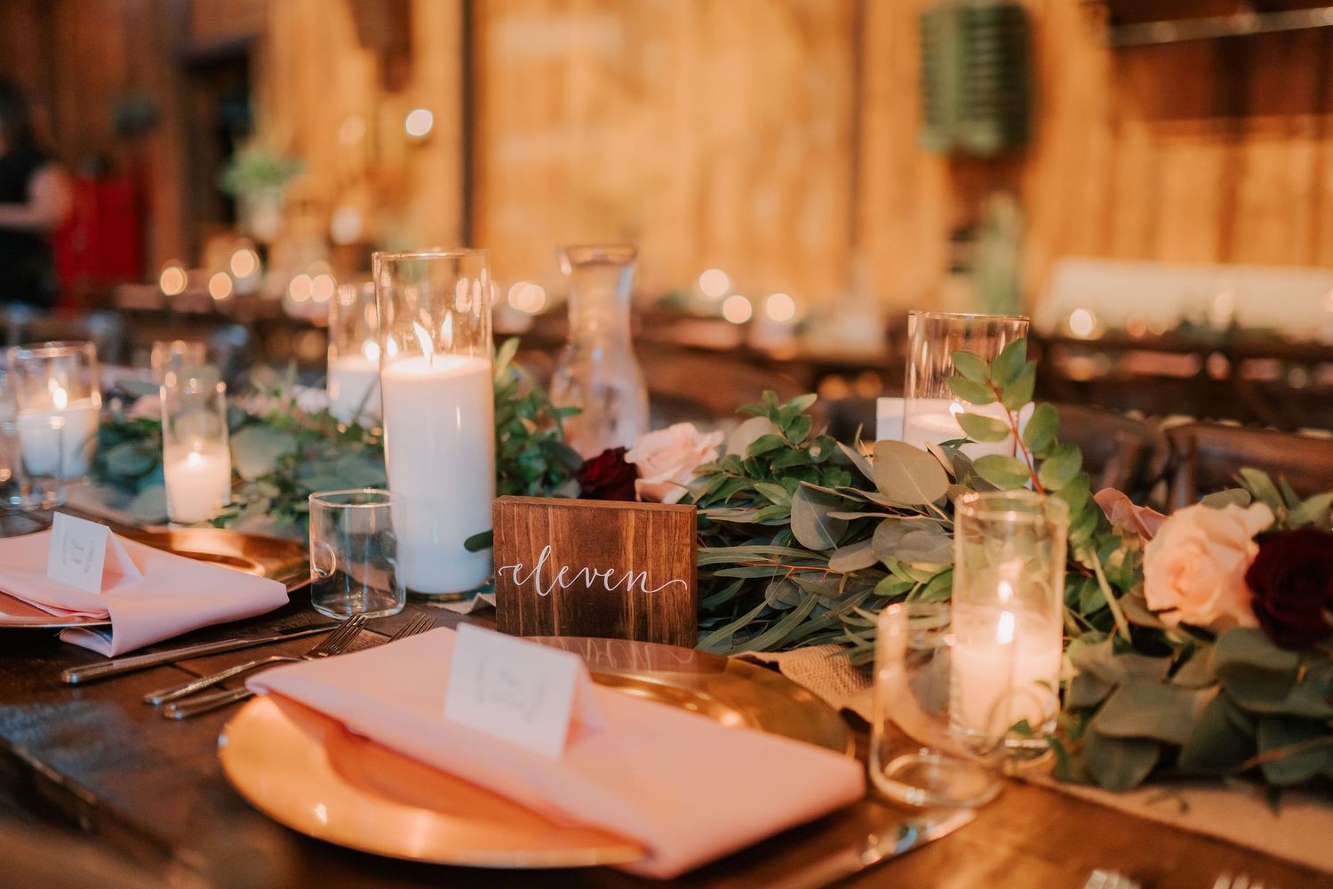 Elegant rustic wedding reception table with candles, greenery runner, gold chargers, blush napkins, and a wooden table number.