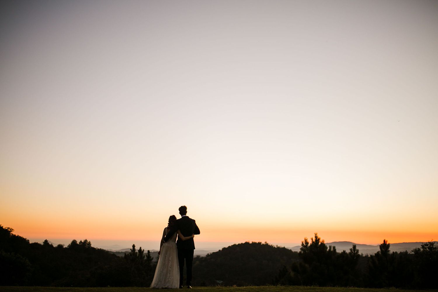 Silhouette of bride and groom embracing at sunset with rolling hills and colorful sky backdrop.