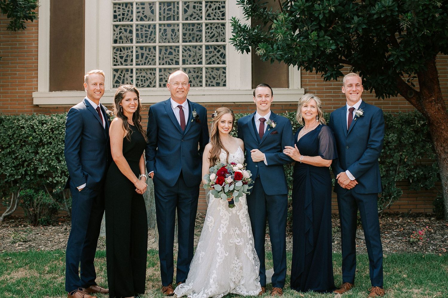 Wedding party poses outdoors in navy attire with bride in white lace gown holding red bouquet before brick building.