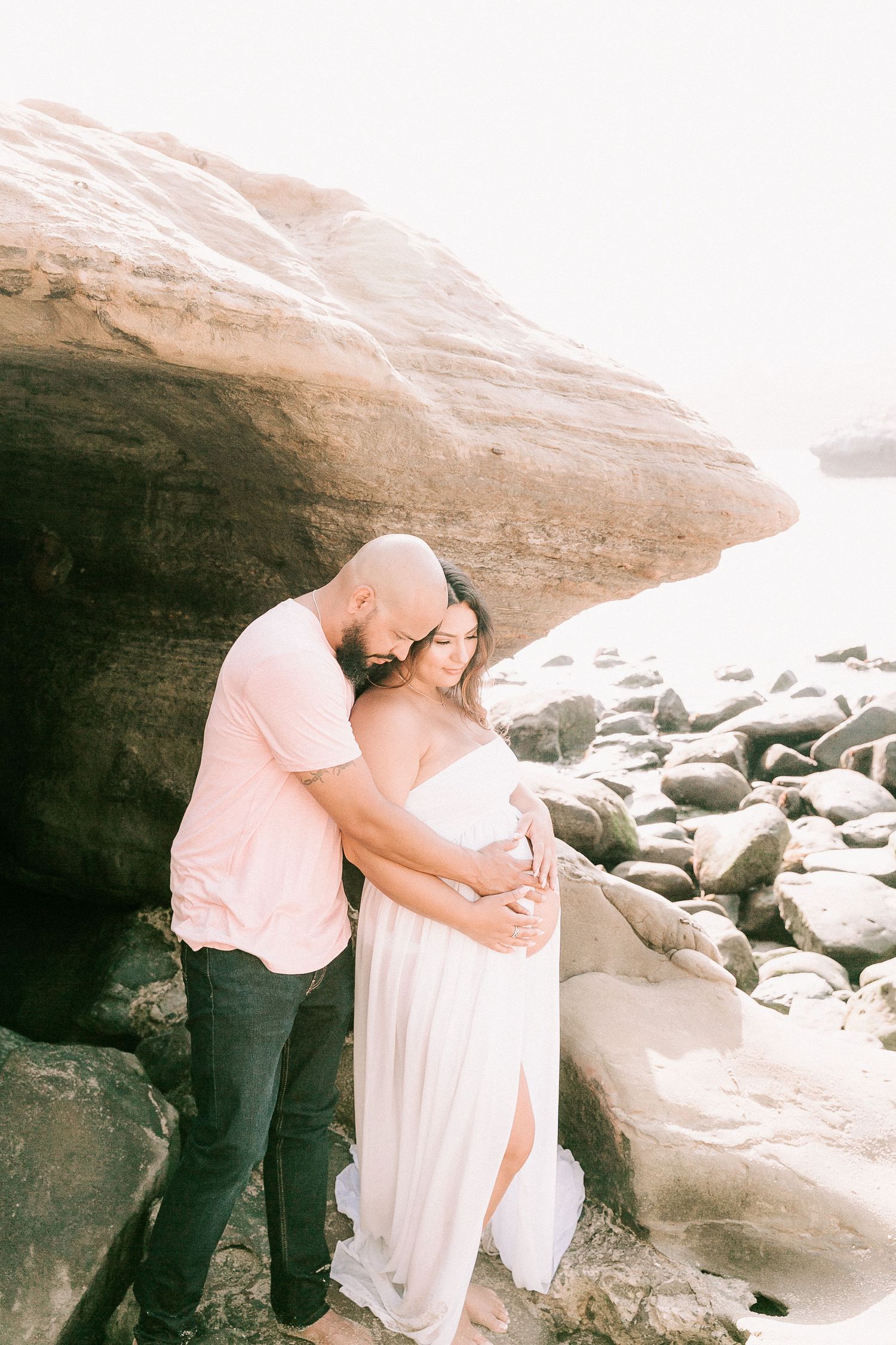 Couple embracing near large rock formation on beach, pregnant woman in white flowing maternity dress.