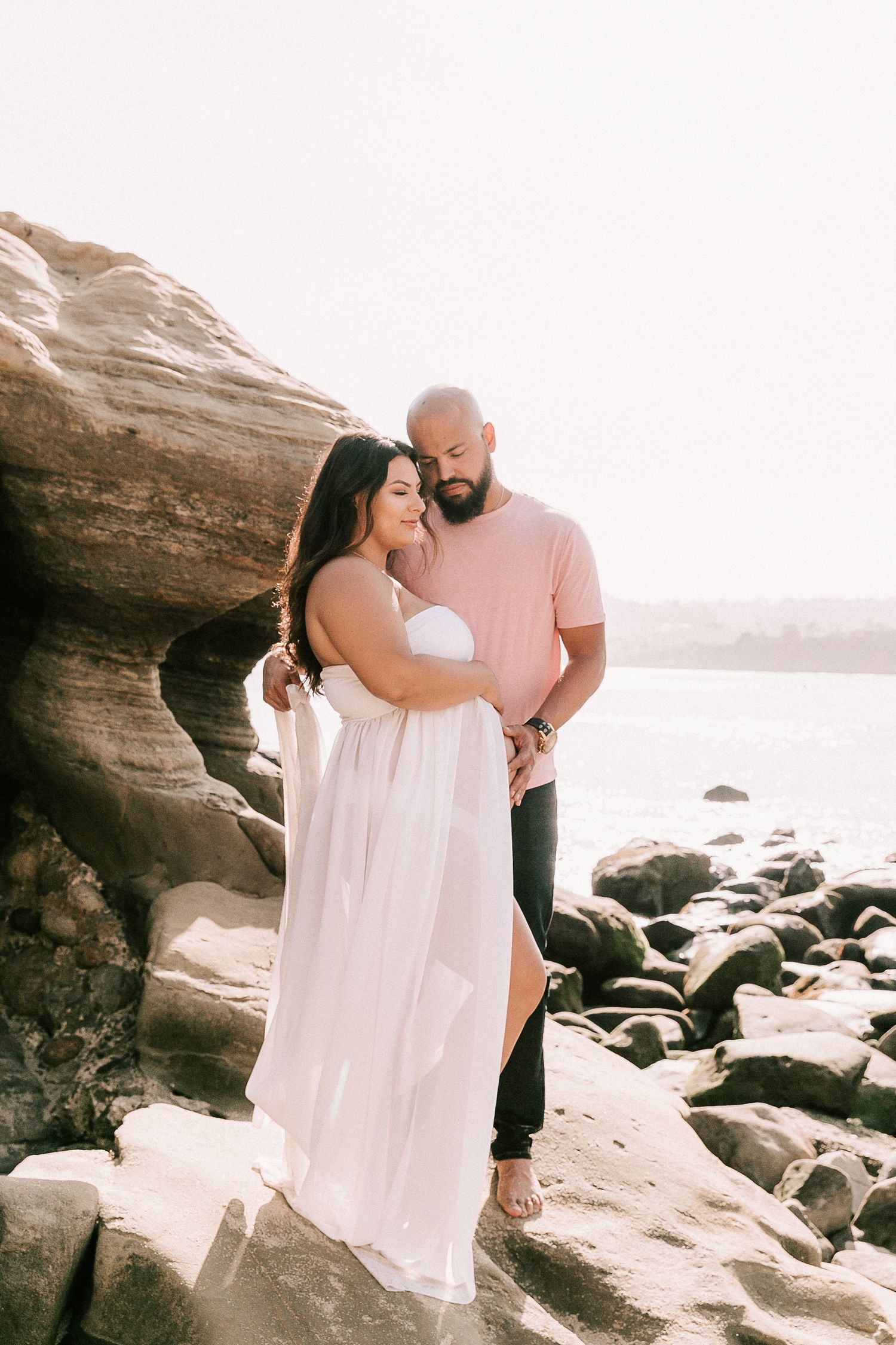 Couple embracing on rocky beach, pregnant woman in white flowing dress, man in pink shirt at golden hour.