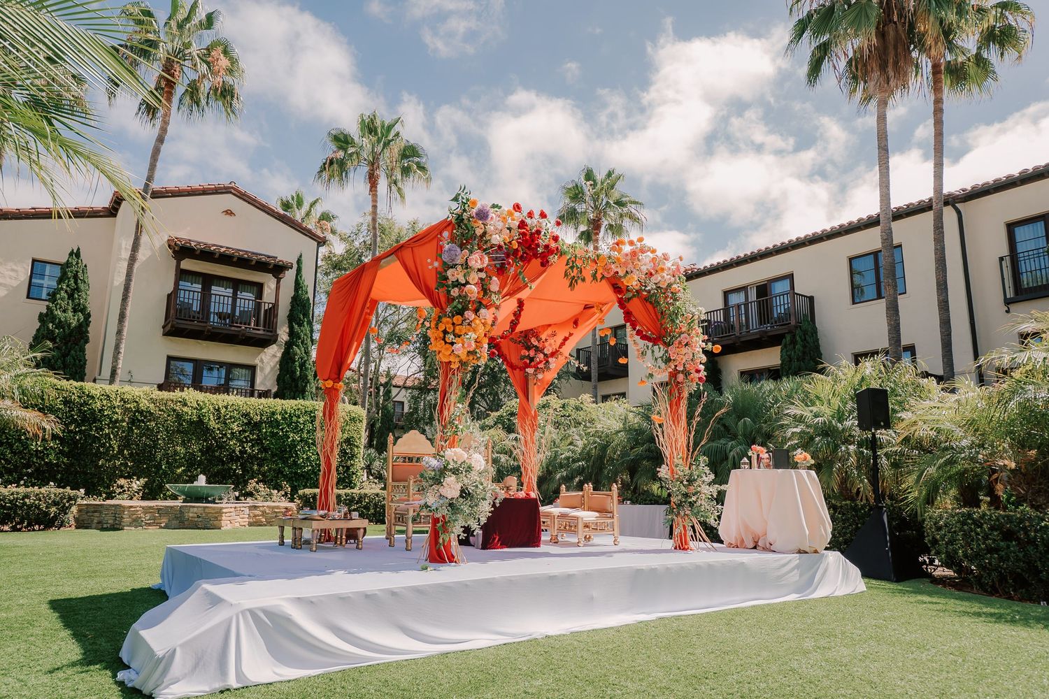 Colorful Indian wedding mandap with orange draping and floral arrangements on outdoor lawn.