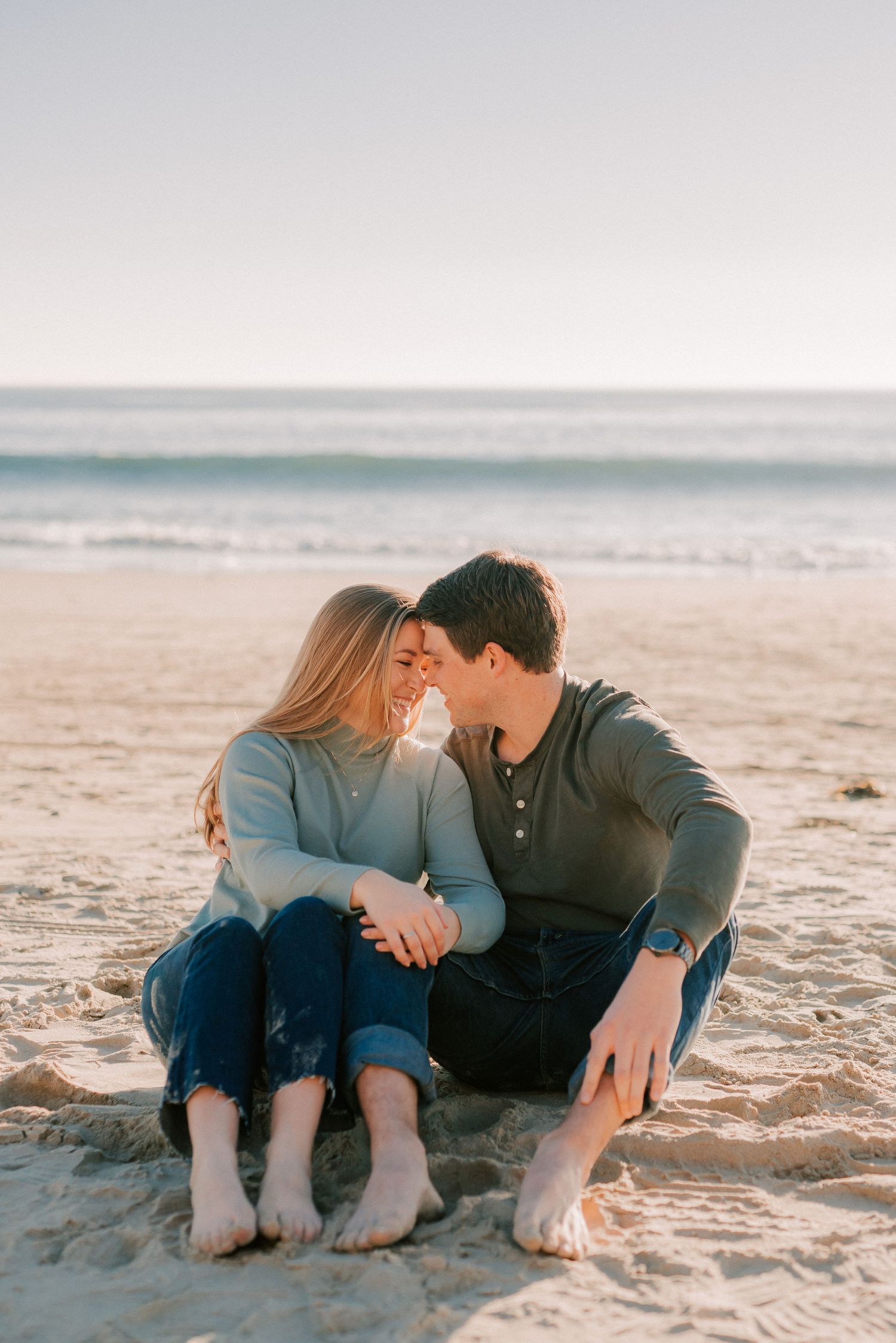 Couple sitting barefoot on sandy beach in warm sunlight sharing a tender forehead touch.