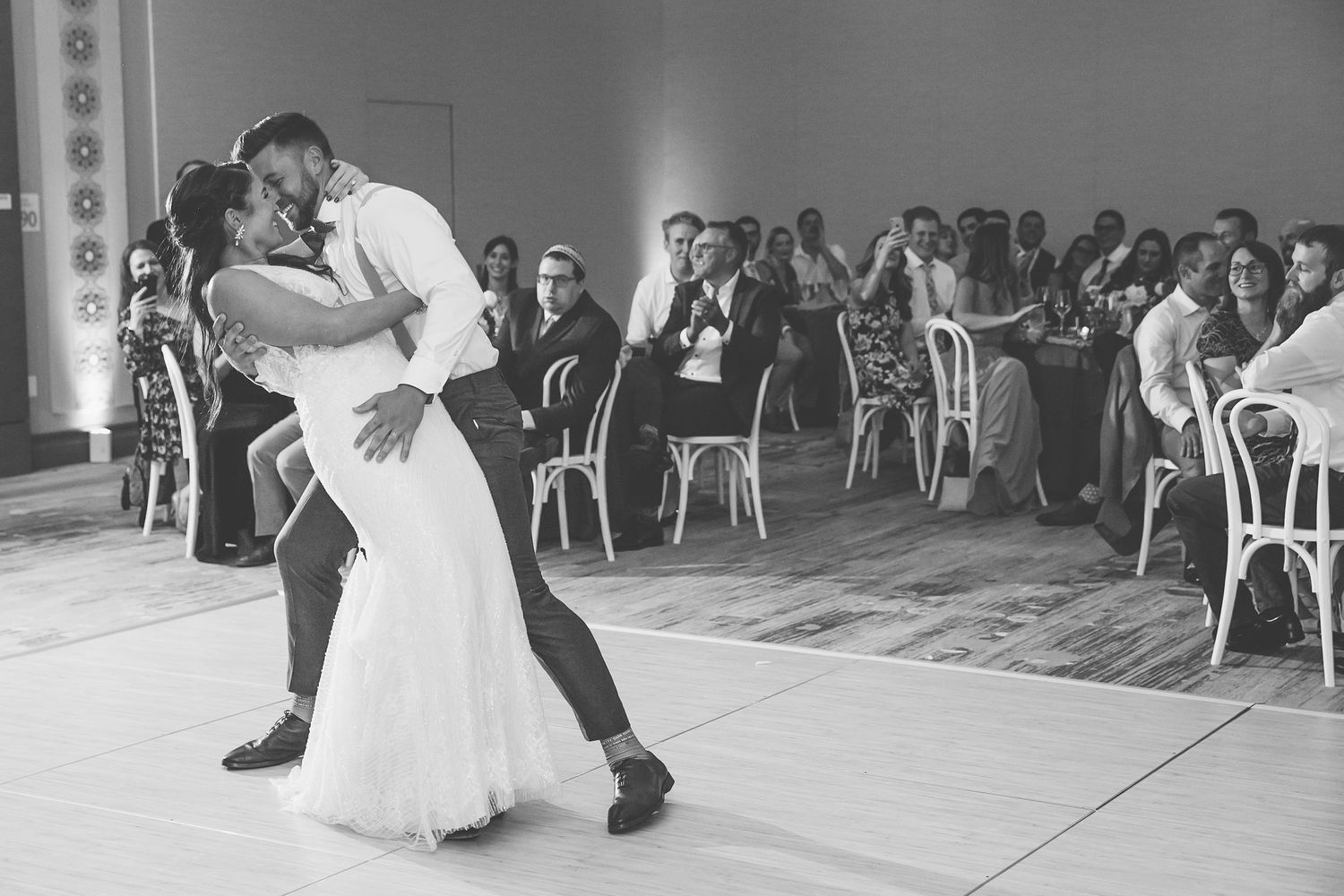 Black and white photo of a couple sharing their first dance dip at a wedding reception with guests seated in background.