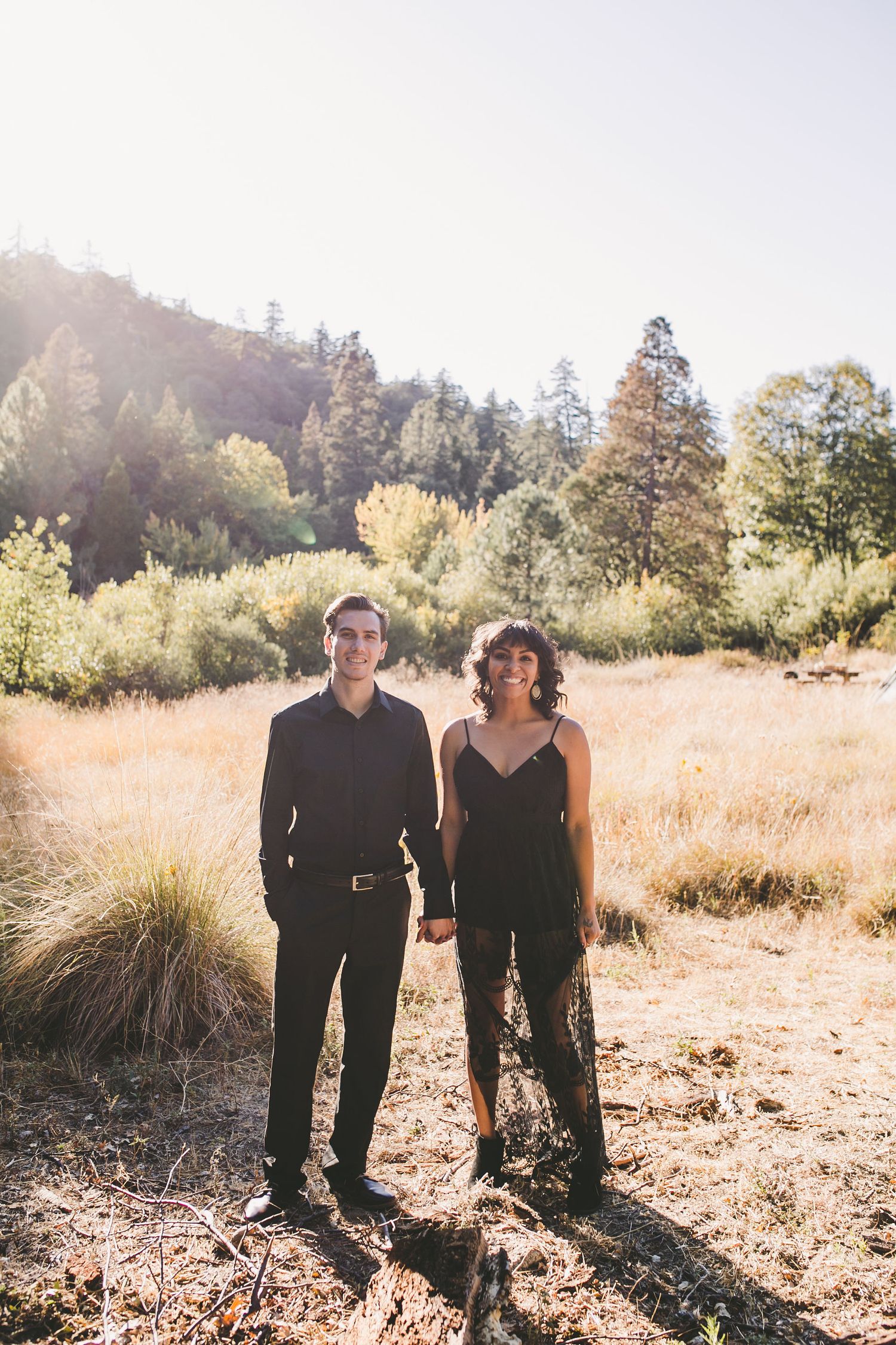 Couple in matching black outfits holding hands in a sunlit meadow with trees and hills in the background.