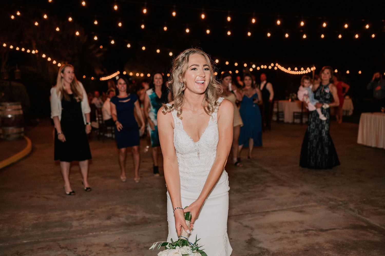 Bride in white gown smiling during bouquet toss at outdoor evening wedding reception with string lights.