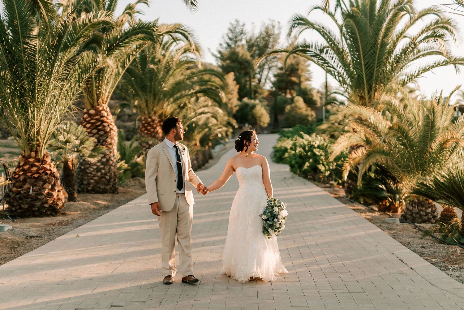 Bride and groom holding hands walking down a palm tree-lined pathway in golden sunset light.
