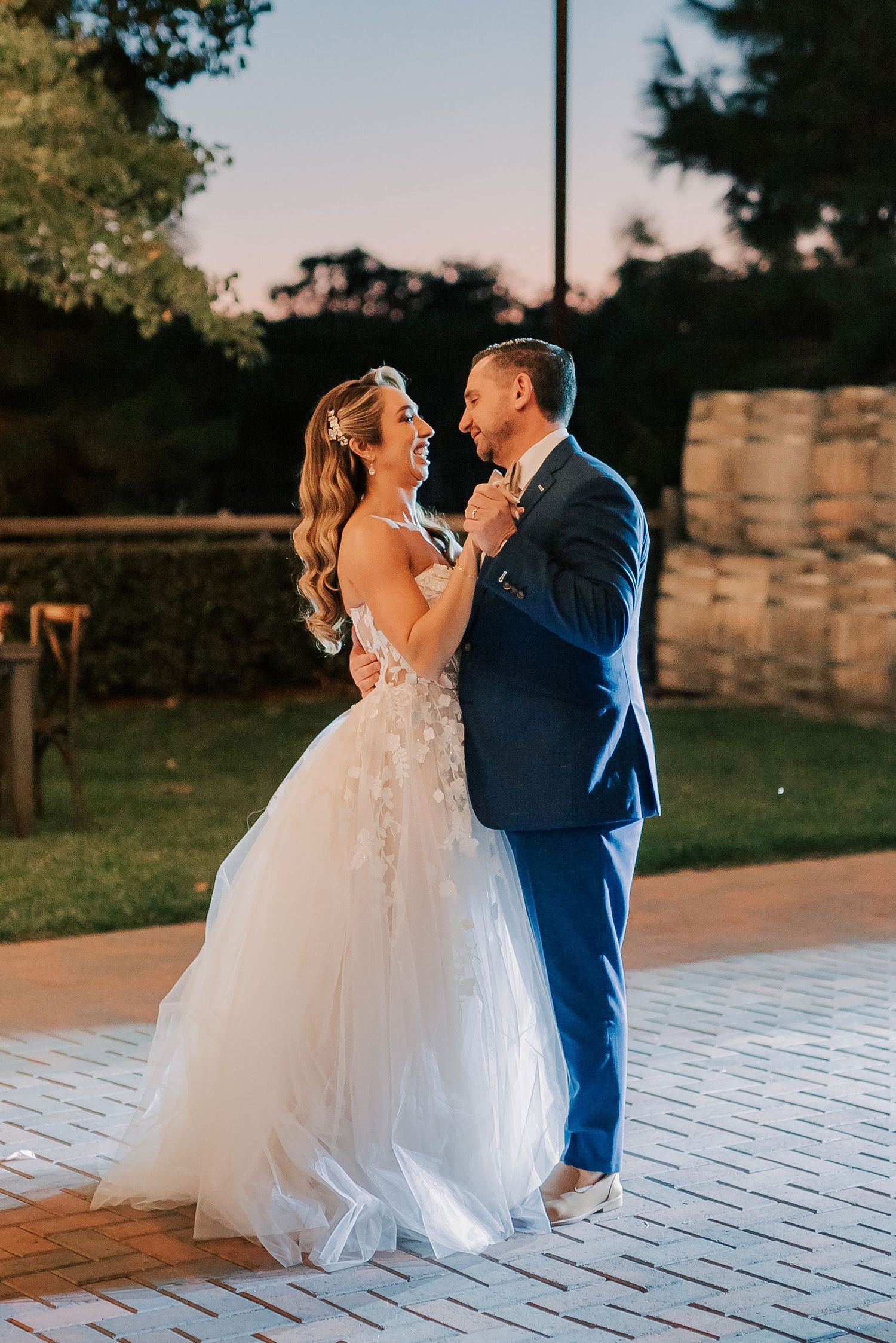 Bride and groom share first dance outdoors at sunset, bride in floral ballgown, groom in navy suit.