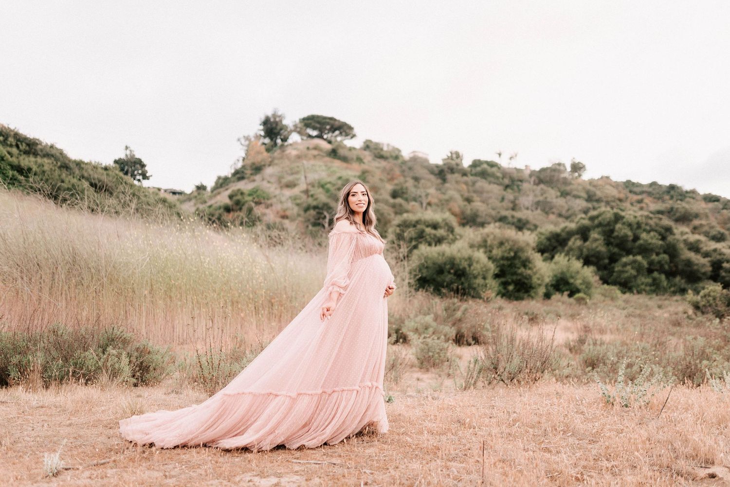 Woman in flowing pink gown poses gracefully in golden field with green hills backdrop.