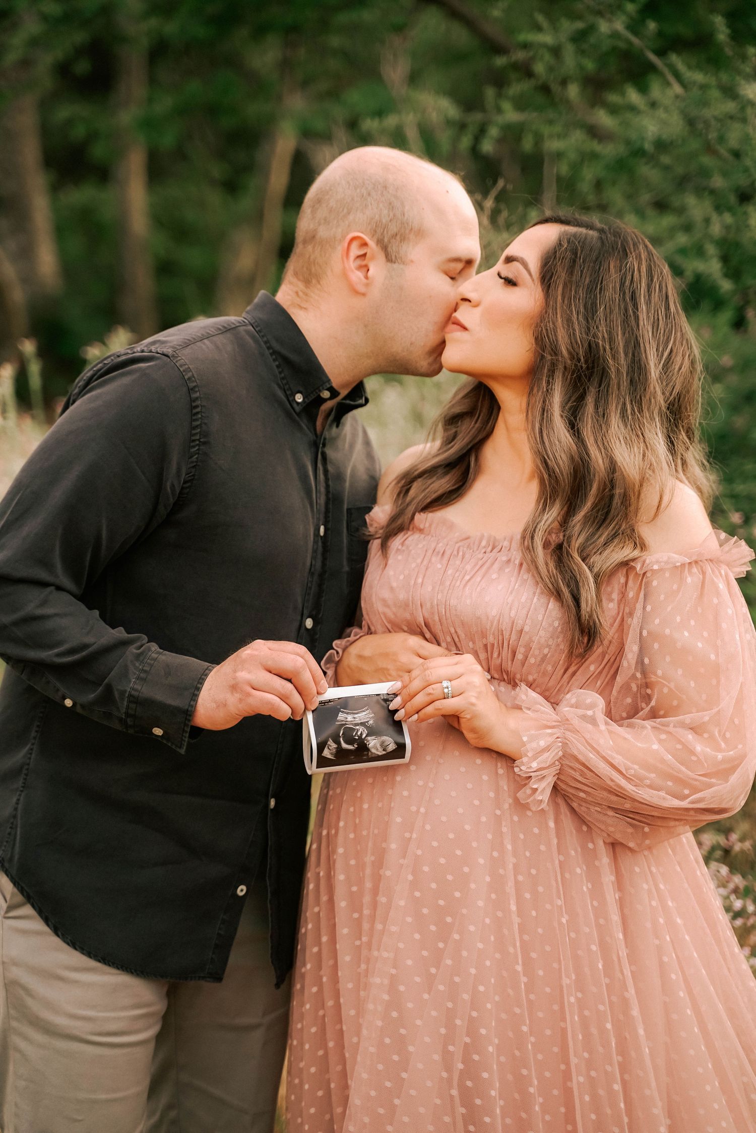 Pregnant couple kissing outdoors, holding ultrasound photo, woman in pink dress, man in black shirt, green trees background.