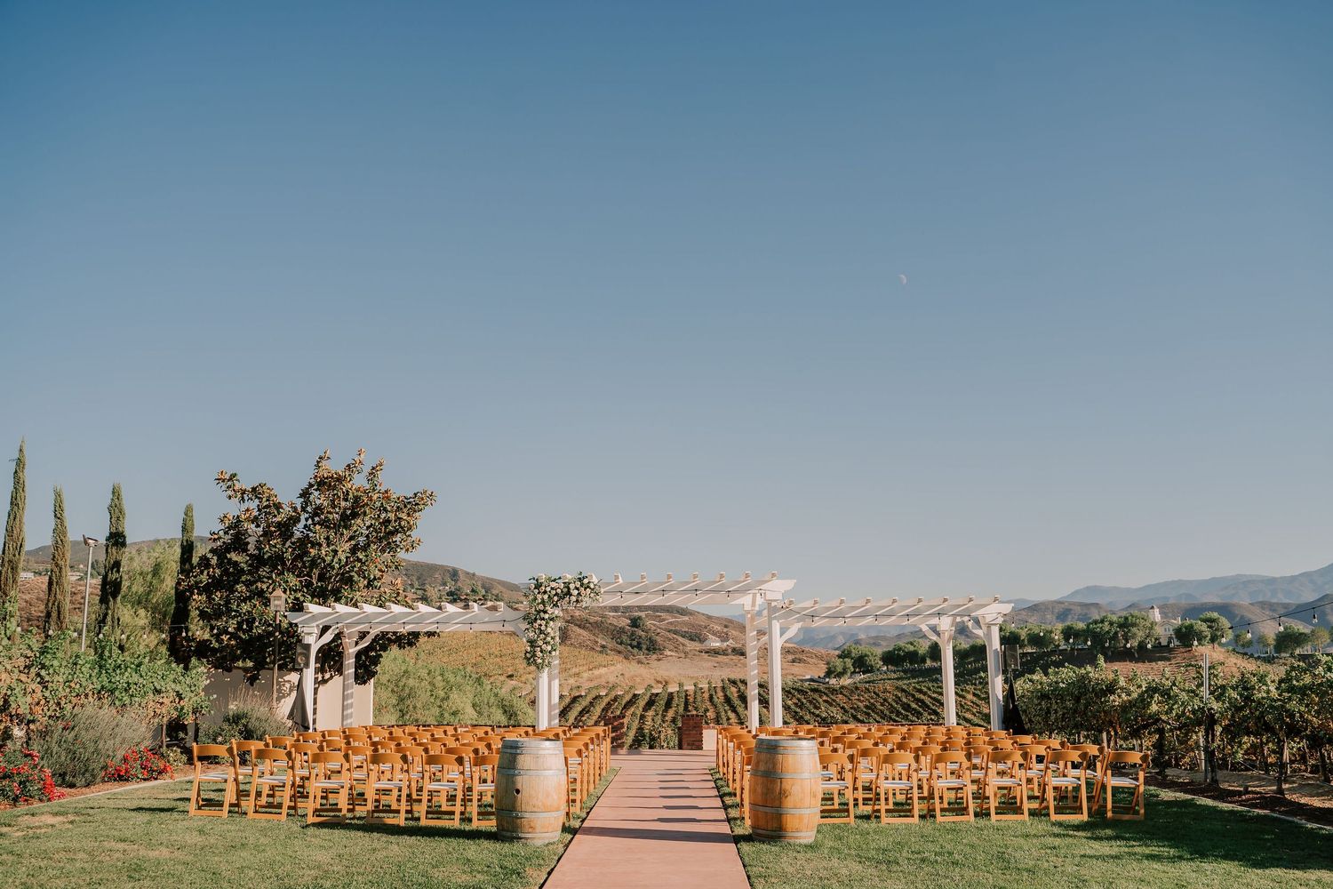 Outdoor vineyard wedding ceremony setup with wooden chairs, barrel aisle markers, white pergola, and rolling hills backdrop.