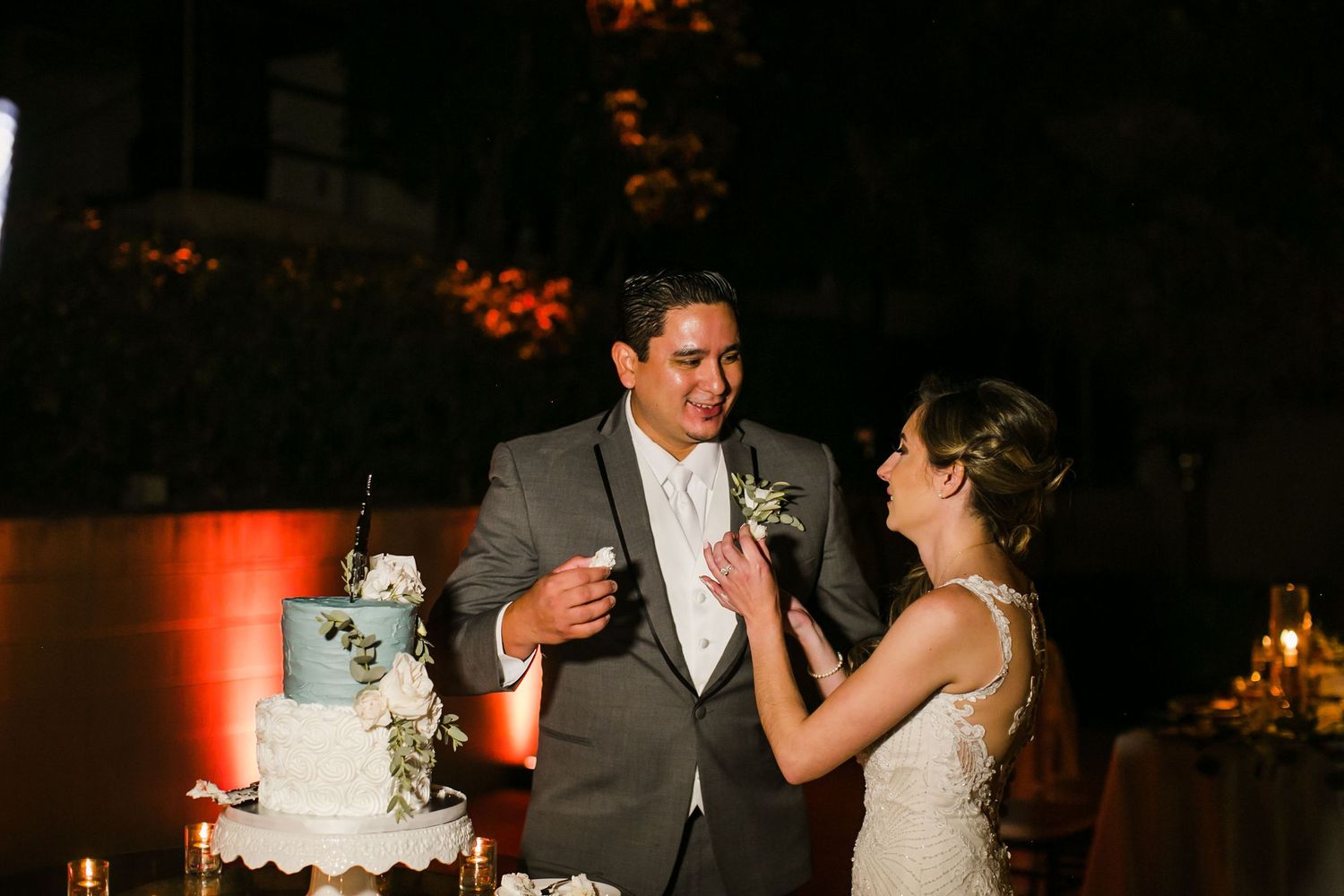 Bride and groom cutting their elegant white wedding cake together at an outdoor evening reception with warm amber lighting.