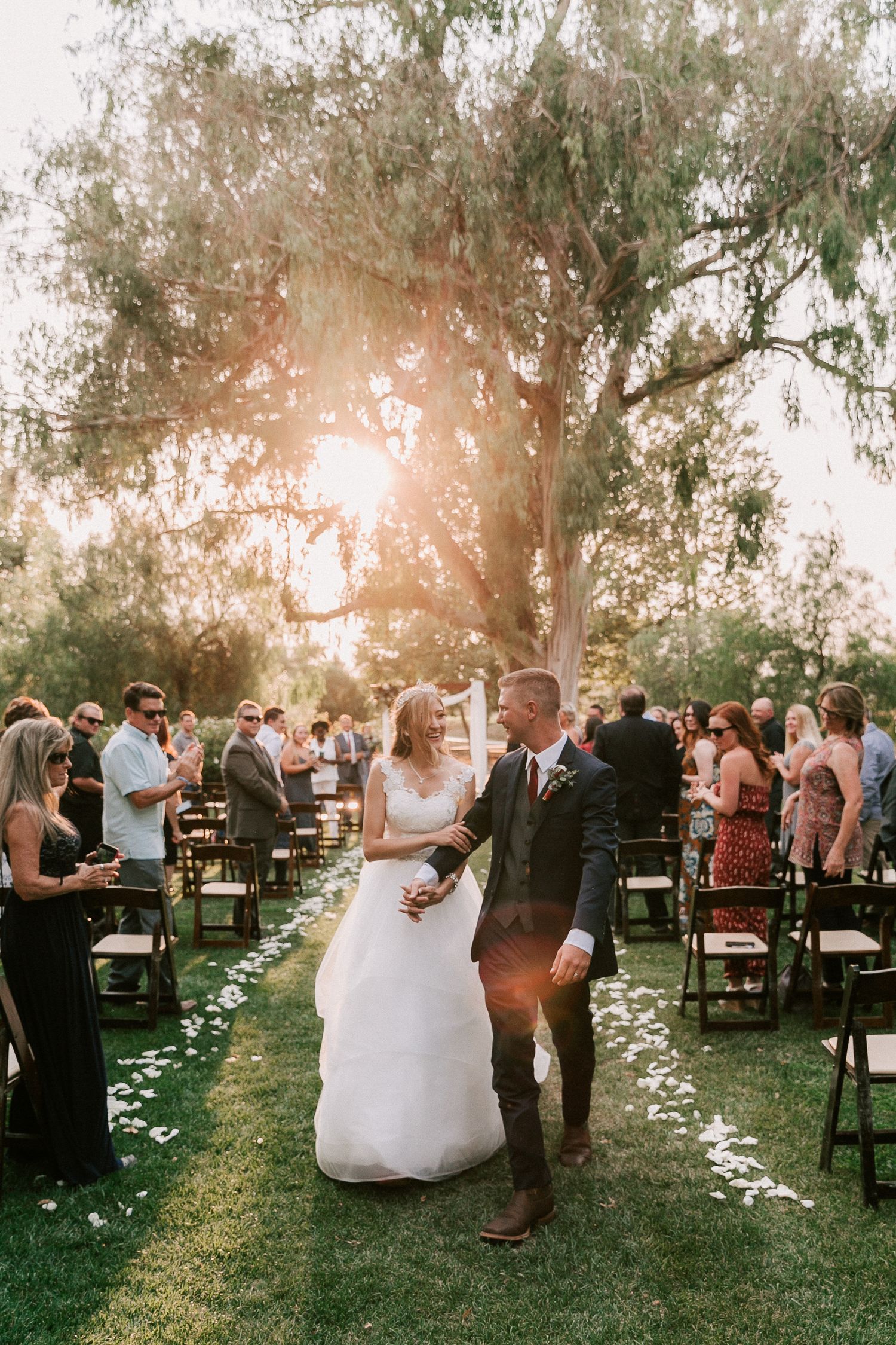 Bride and groom walk down outdoor aisle after wedding ceremony, surrounded by guests under a sunlit tree.