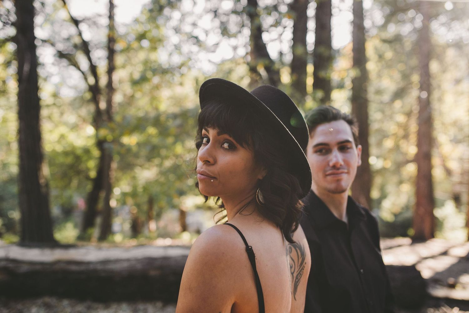 A young woman in a black hat and a man stand among tall trees in a sunlit forest setting.