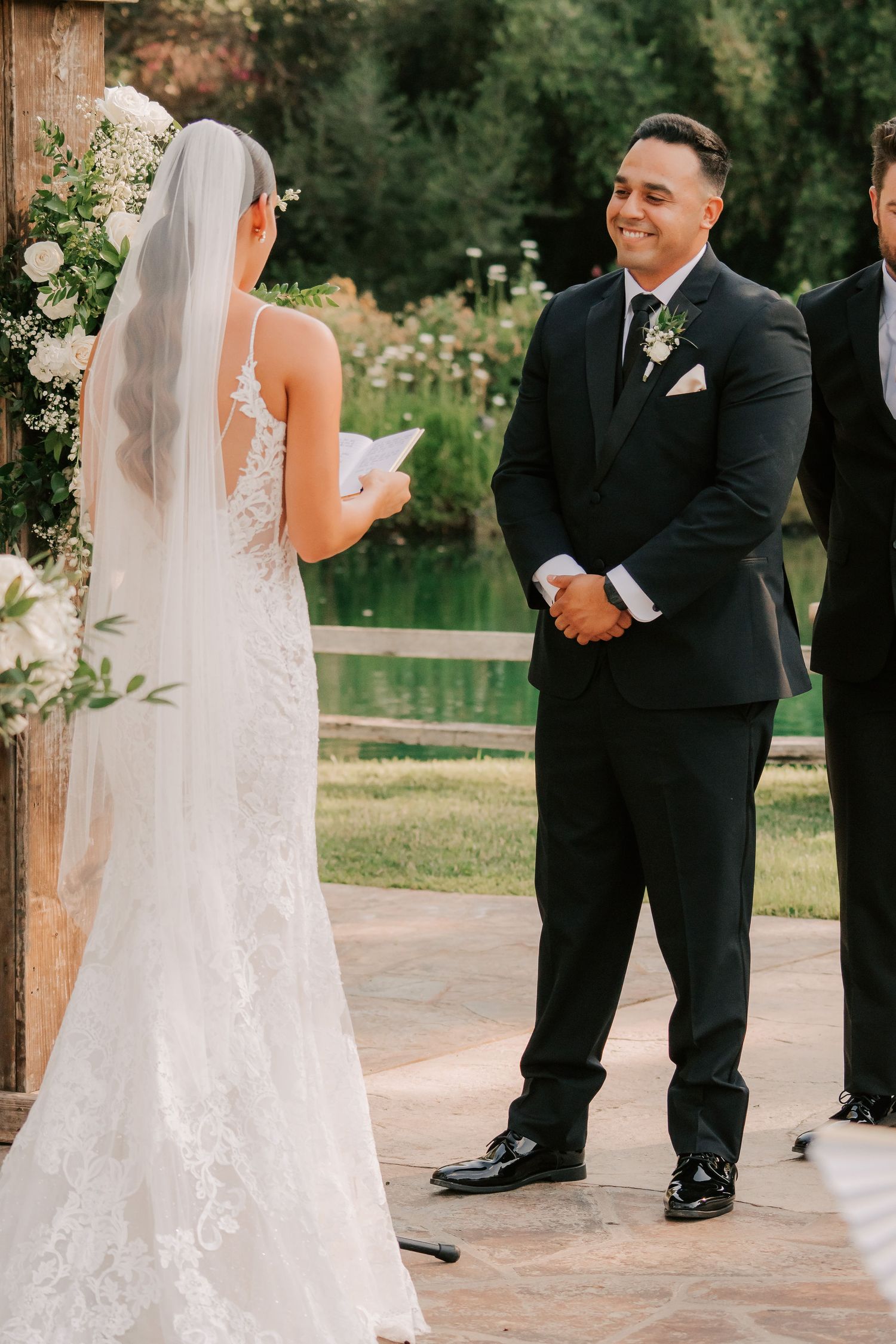 Groom smiling at bride reading vows during outdoor wedding ceremony with floral arch and pond backdrop.