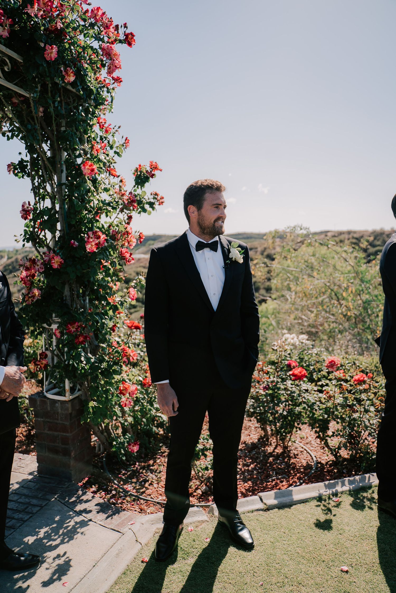 Groom in black tuxedo with bow tie stands at outdoor wedding altar adorned with red climbing roses.