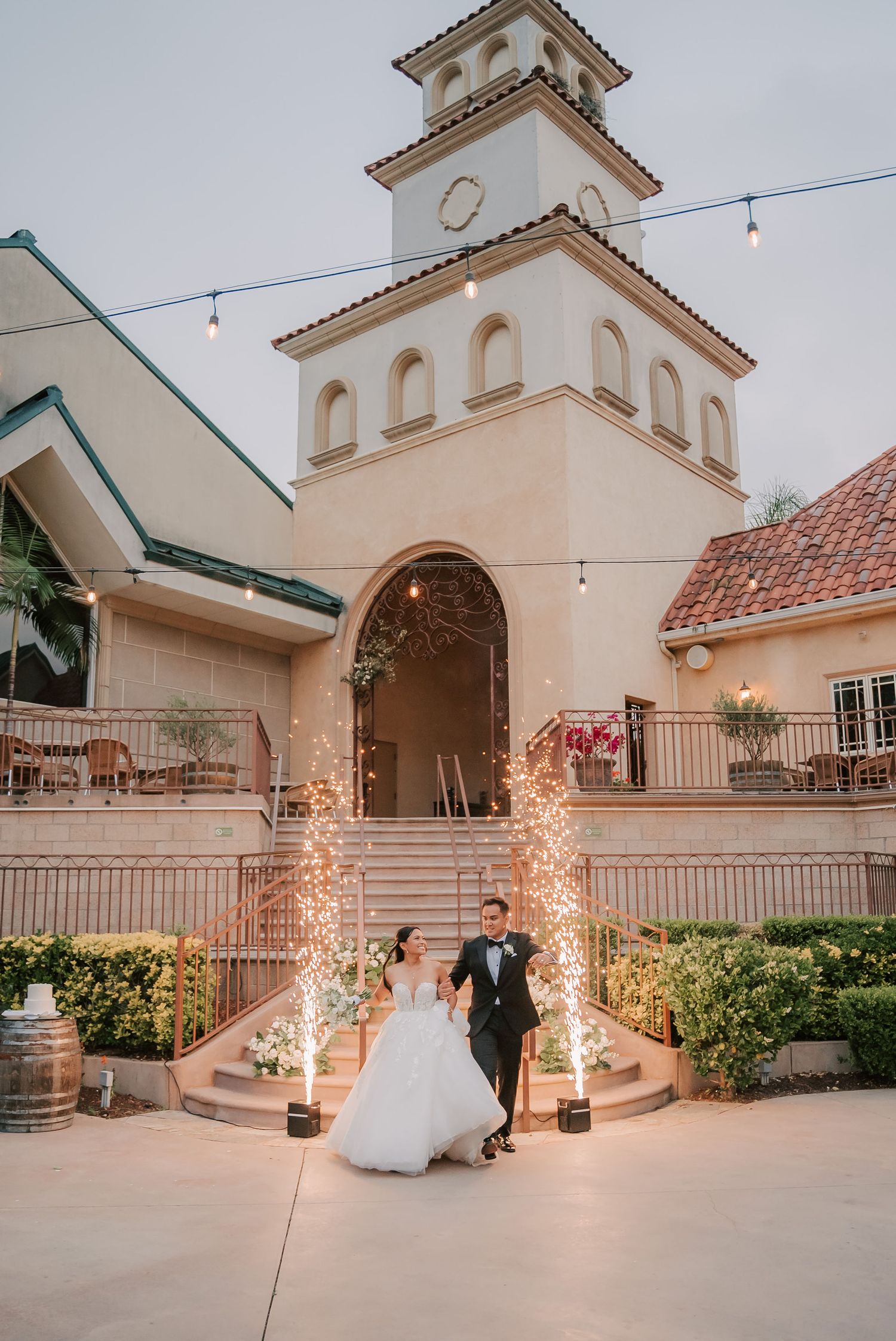 Bride and groom exit wedding venue with sparkling fireworks fountains in front of elegant Spanish-style bell tower at dusk.