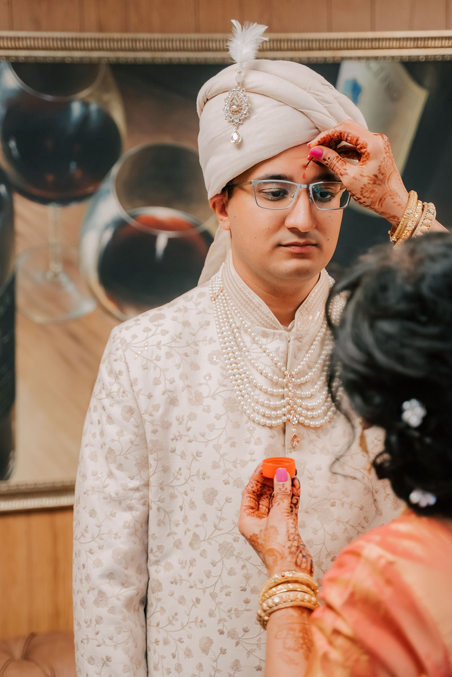 Indian groom in white sherwani and safa turban receives tilak blessing from woman with mehndi hands.