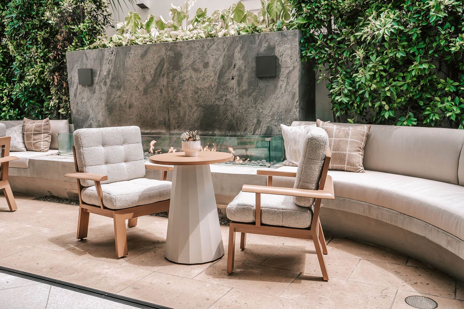 Elegant outdoor patio with curved white sofa, wooden armchairs, round table, and lush green living wall backdrop.