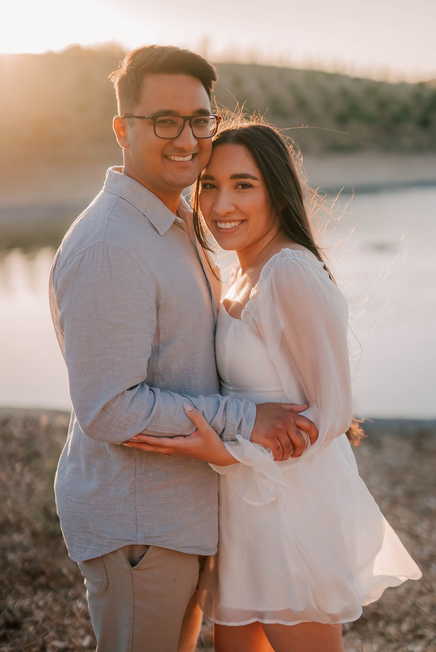 Couple embracing and smiling at sunset near a lake, woman in white dress and man in light blue shirt.