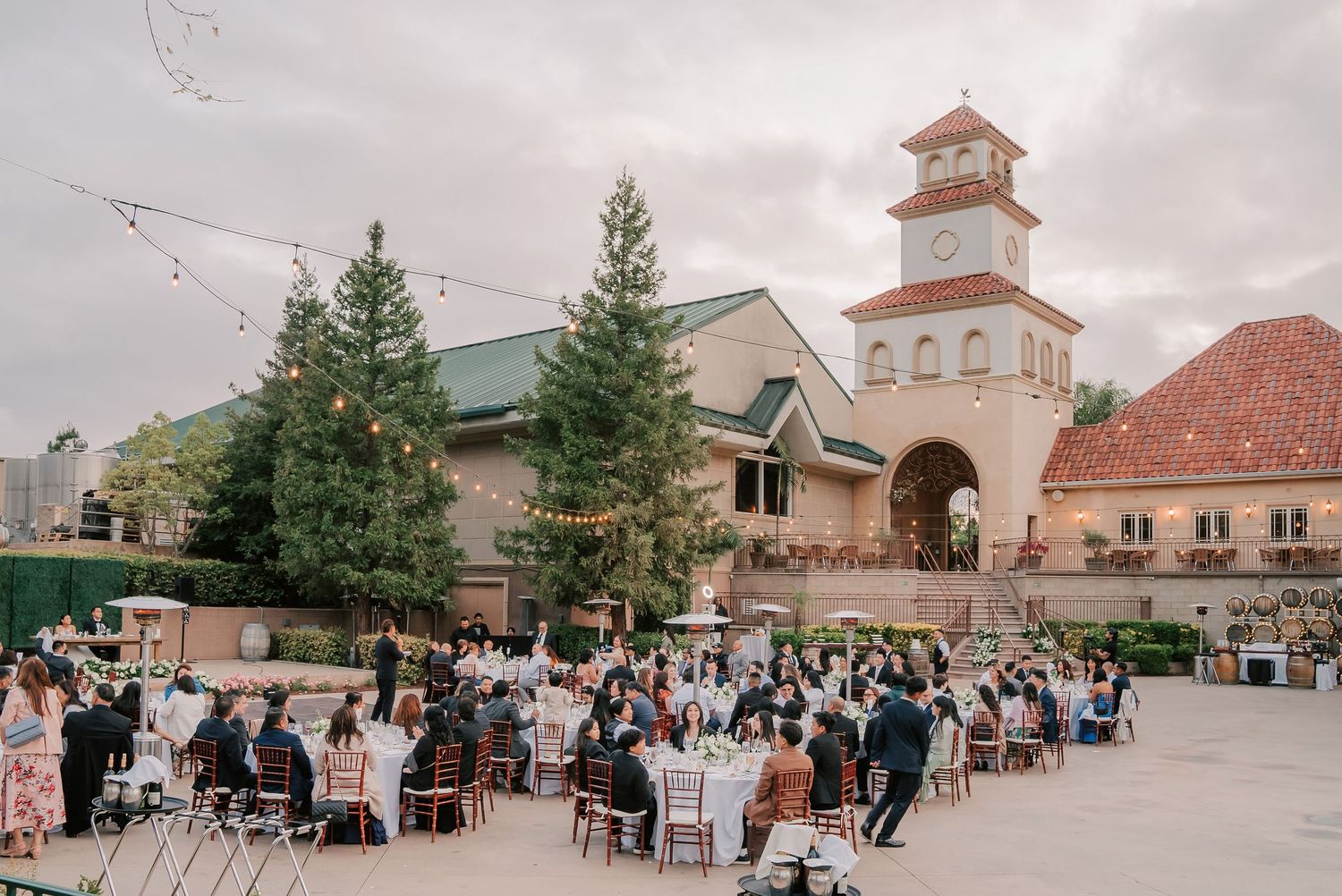 Outdoor wedding reception at a Spanish-style venue with string lights, tall trees, and guests seated at elegant tables.