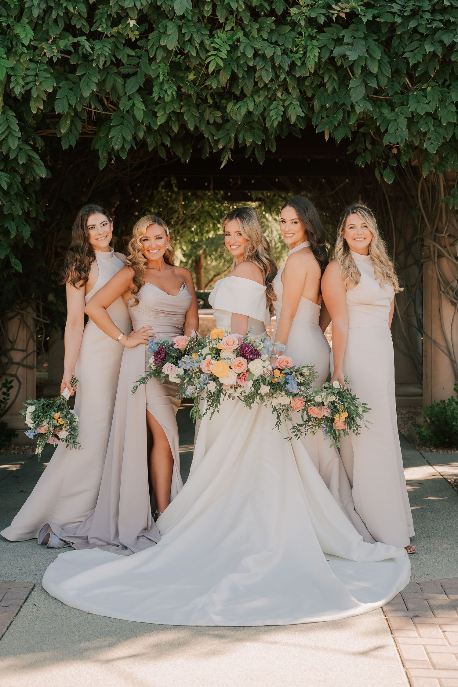 Bride in white gown with bridesmaids in taupe dresses holding colorful bouquets outdoors under lush green ivy.