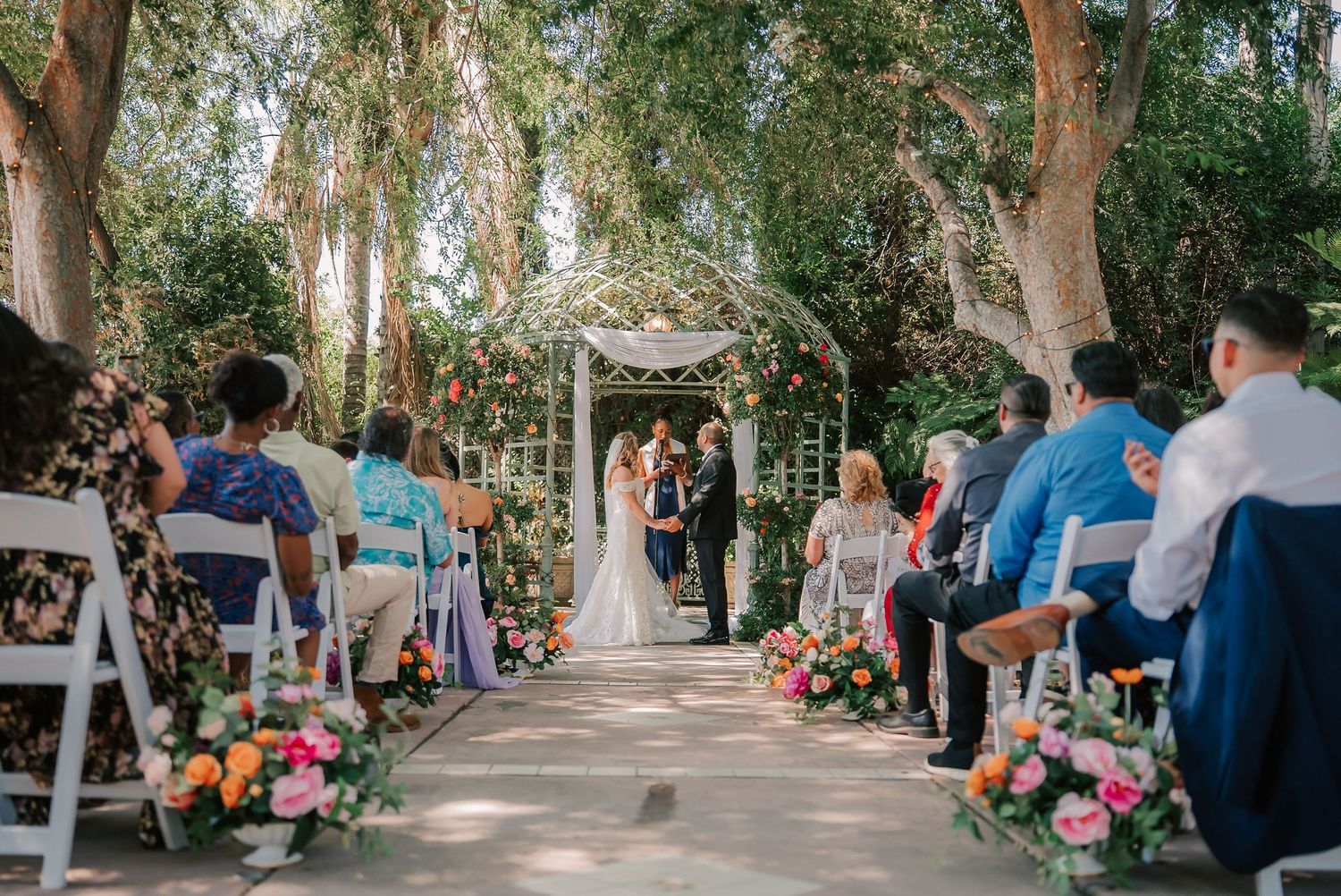 Outdoor wedding ceremony under floral arch with guests seated on white chairs along colorful flower-lined aisle.