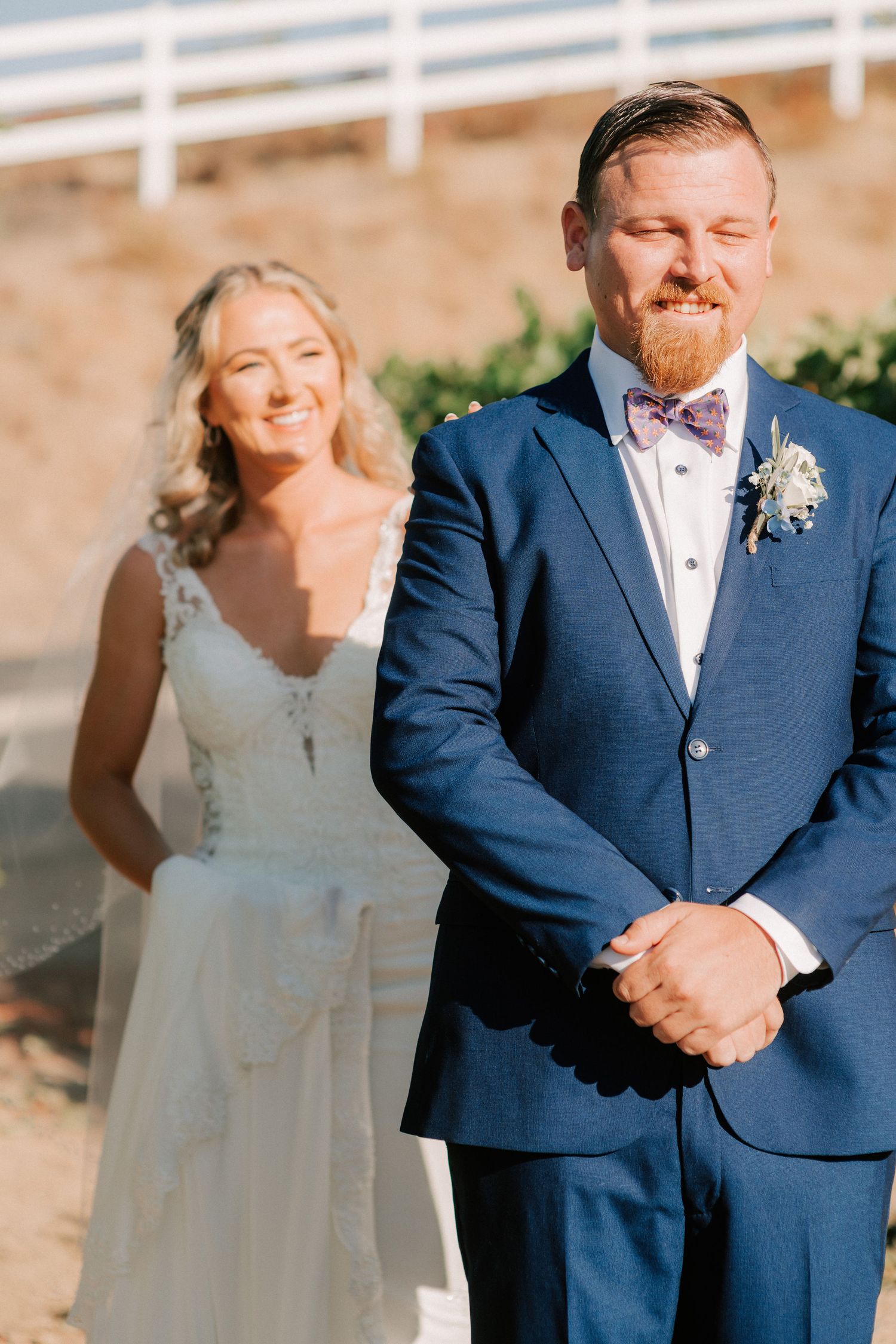 Bride in lace gown smiling behind groom in navy suit with bow tie during outdoor wedding first look.