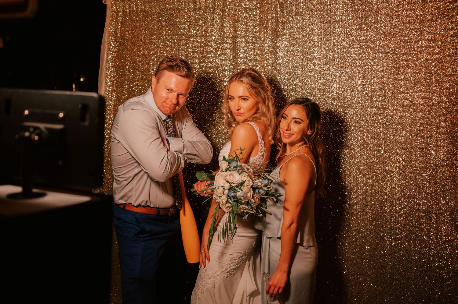 Bride in gray gown with bouquet poses with groom and bridesmaid in front of gold sequin backdrop.