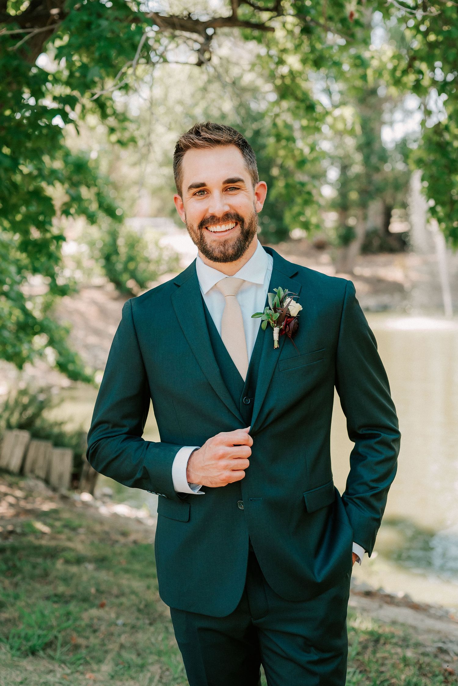 Smiling groom in dark teal suit with boutonniere and tan tie poses outdoors near a pond surrounded by green trees.